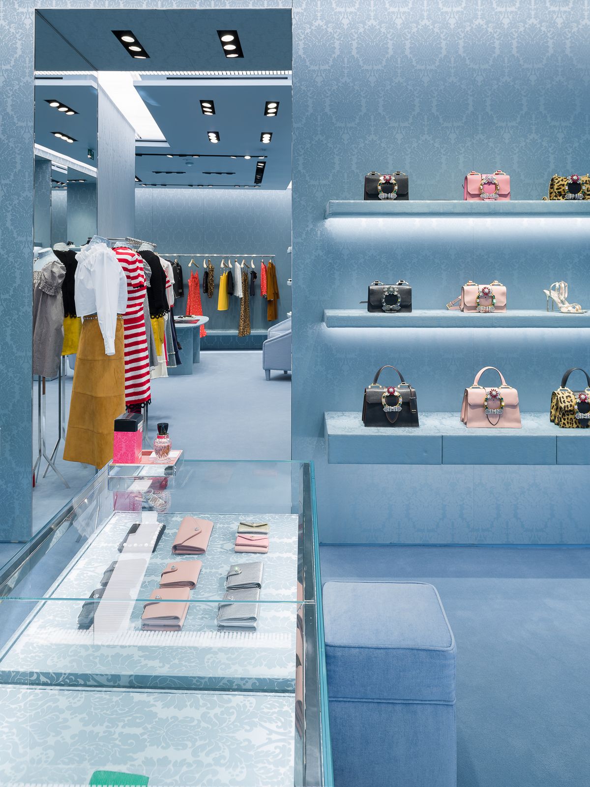 Interior de tienda Miu Miu mostrando paredes estampadas azules, mostradores de exhibición de vidrio con accesorios y percheros de ropa con prendas coloridas.