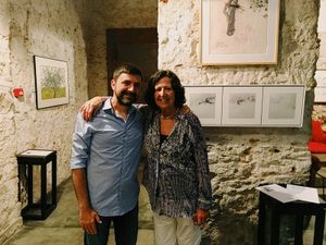 Dos personas de pie y sonriendo en una galería de arte con obras de arte enmarcadas en una pared de piedra en el fondo.