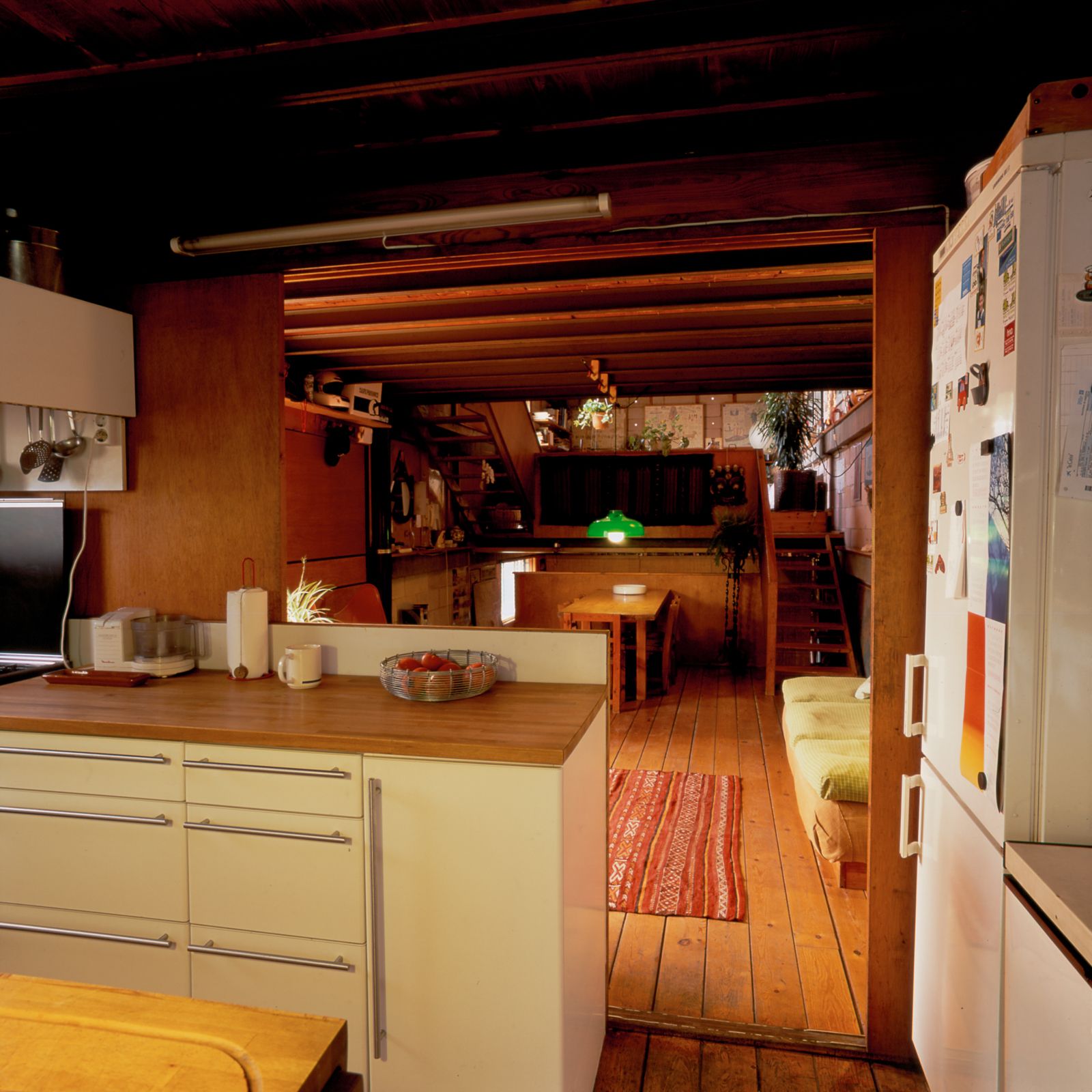 Cocina acogedora con suelo y techo de madera, armarios blancos, que conduce a un comedor con una alfombra colorida, una mesa de madera, lámpara colgante verde y una escalera de madera al aire libre en el fondo.
