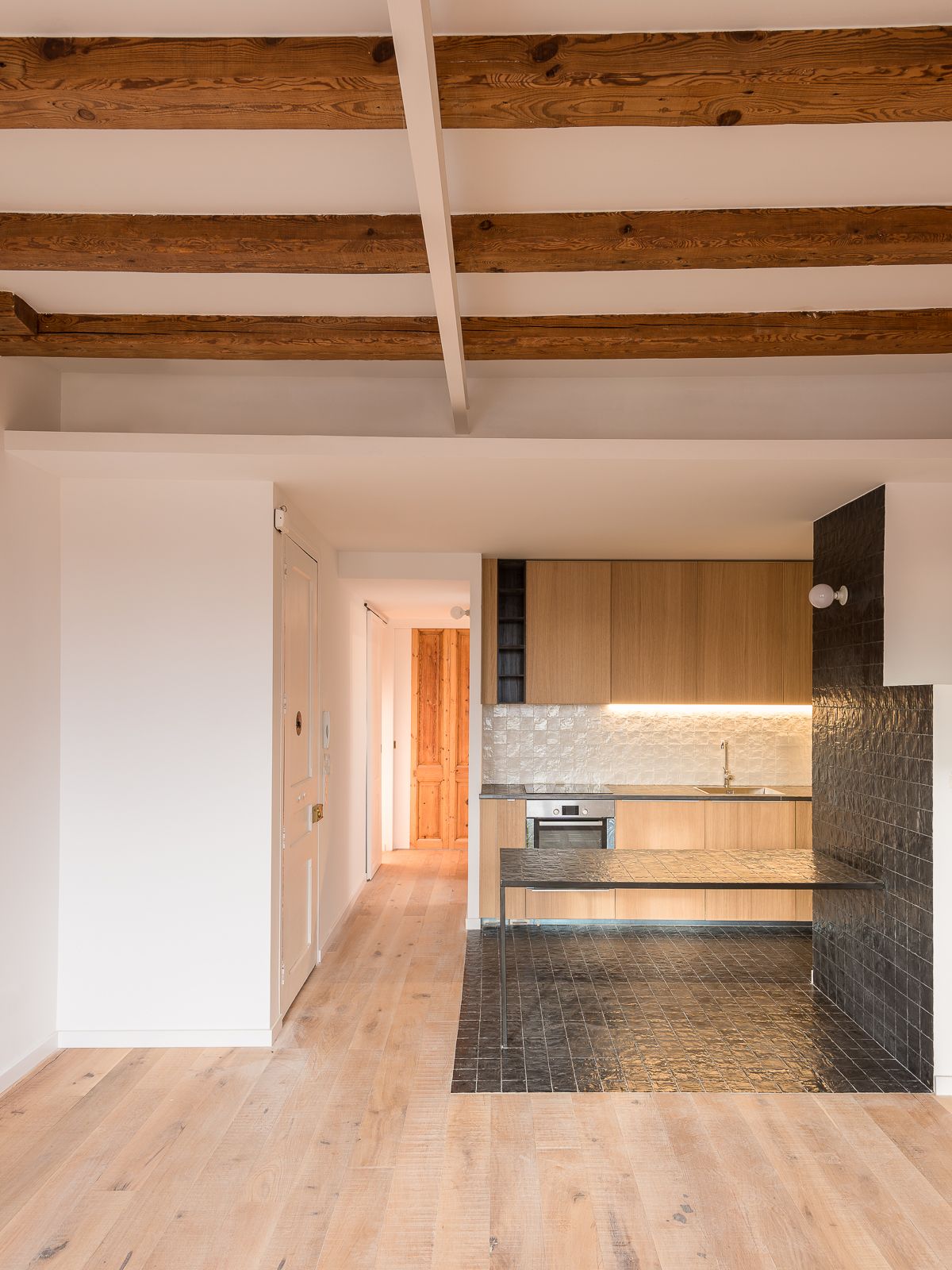 Cocina abierta con armarios de madera, encimera de baldosas negras y vigas de madera en el techo.