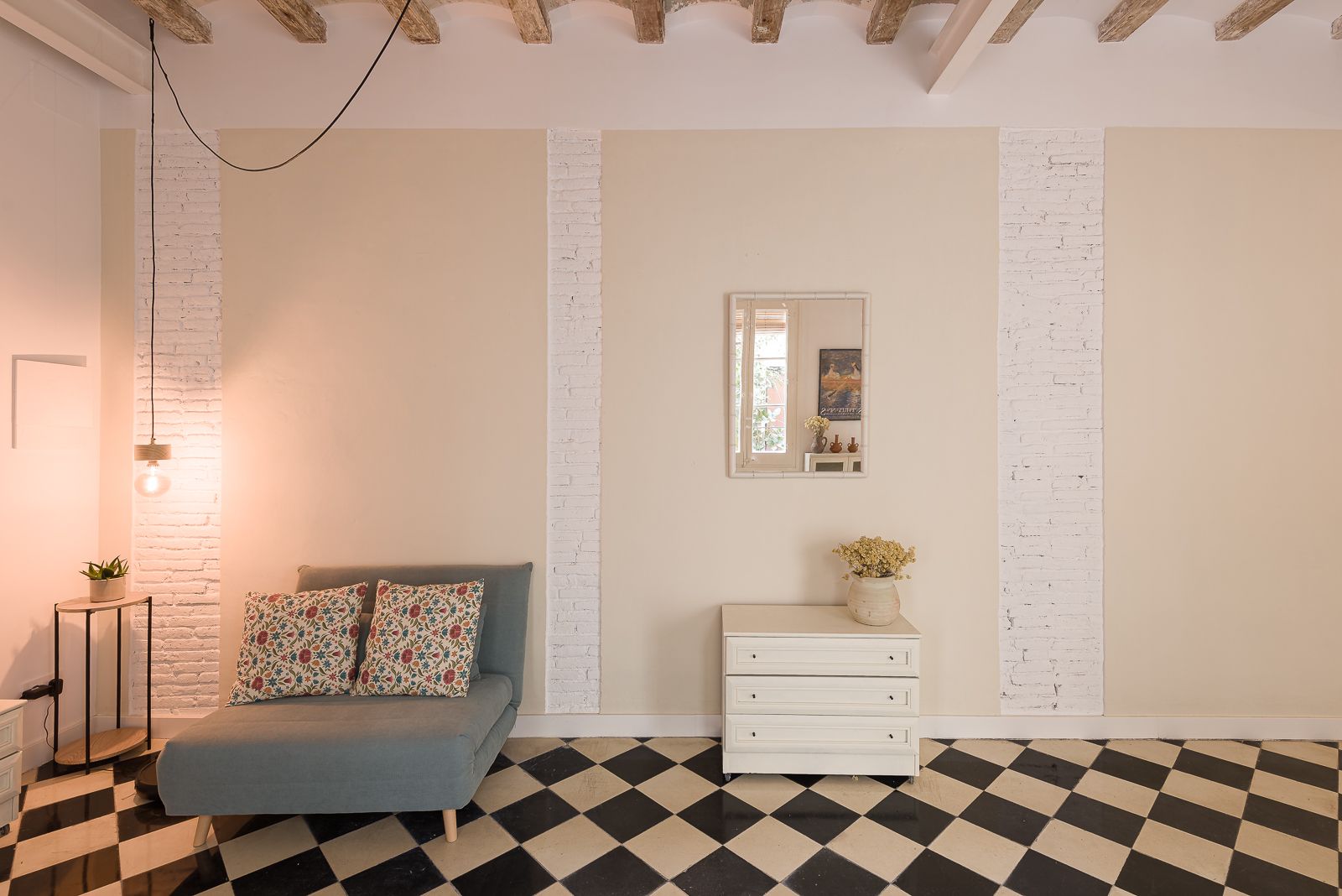 Cozy corner with a blue armchair, floral pillows, a small table, a white dresser, a square mirror on the wall reflecting the door onto the patio and checkered floor tiles.