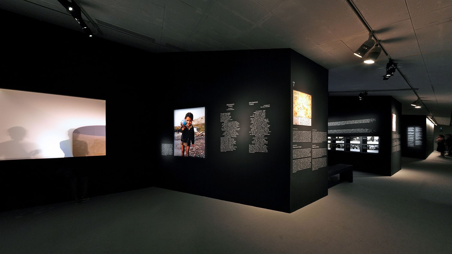 * Vista de ángulo ancho de una sala de exposiciones de un museo moderno con paneles expositivos en negro mate con bloques de texto, una fotografía iluminada de un niño, una pantalla de proyección brillante a la izquierda y un recorrido que continúa al fondo.