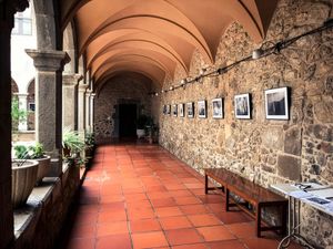 Una fila de fotografías enmarcadas expuestas en una pared de piedra en el corredor cubierto de un claustro con arcos, plantas en macetas y bancos.