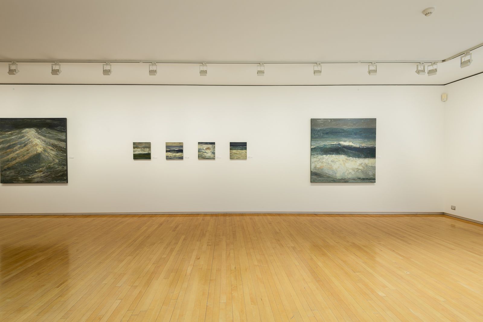Galería de arte con suelos de madera y paredes blancas, con una serie de pinturas de paisajes marinos de diferentes tamaños exhibidas en fila.