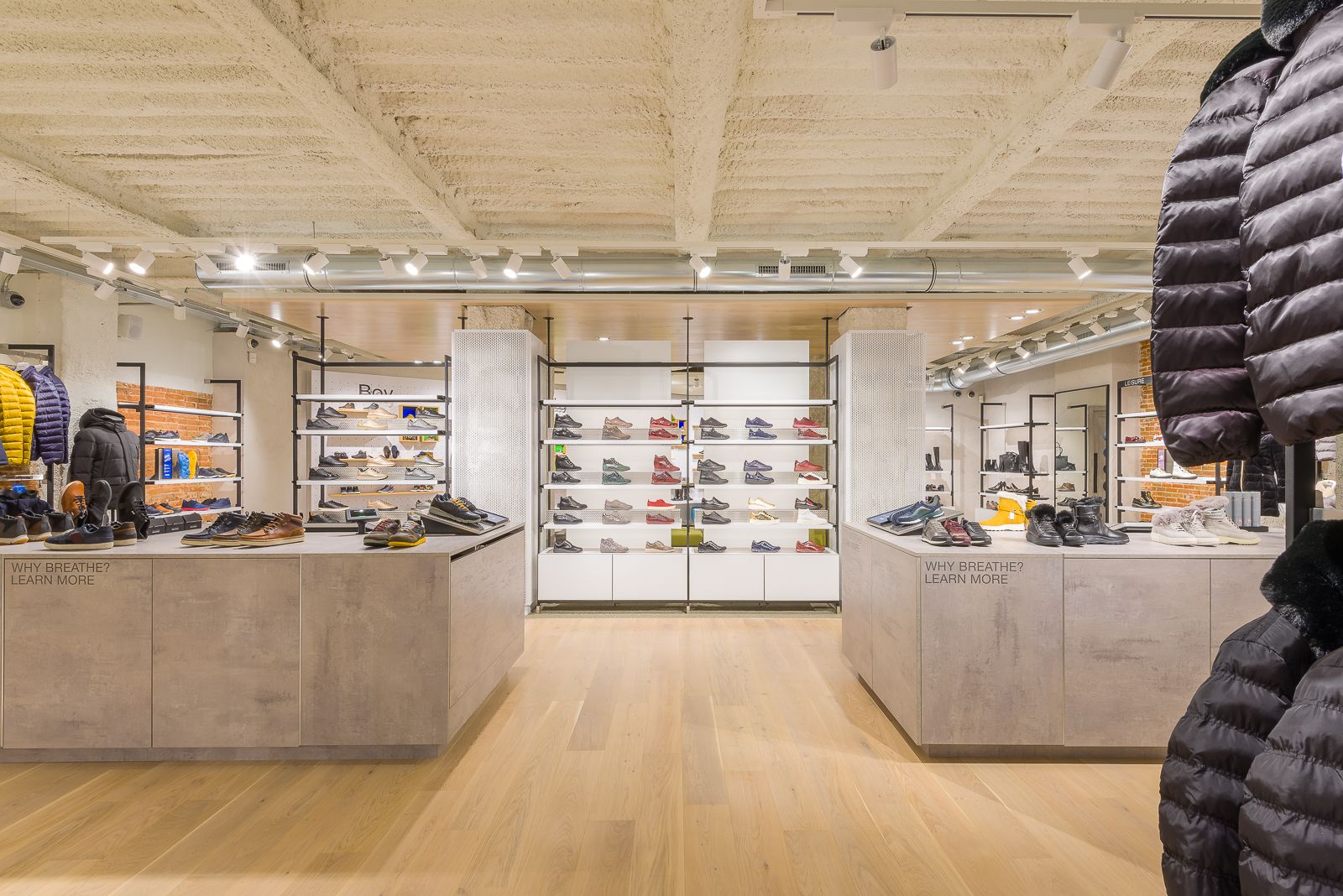 Interior de la tienda Geox, con estantes llenos de varios estilos de zapatos y chaquetas de invierno, con un telón de fondo de ladrillo expuesto e iluminación moderna.