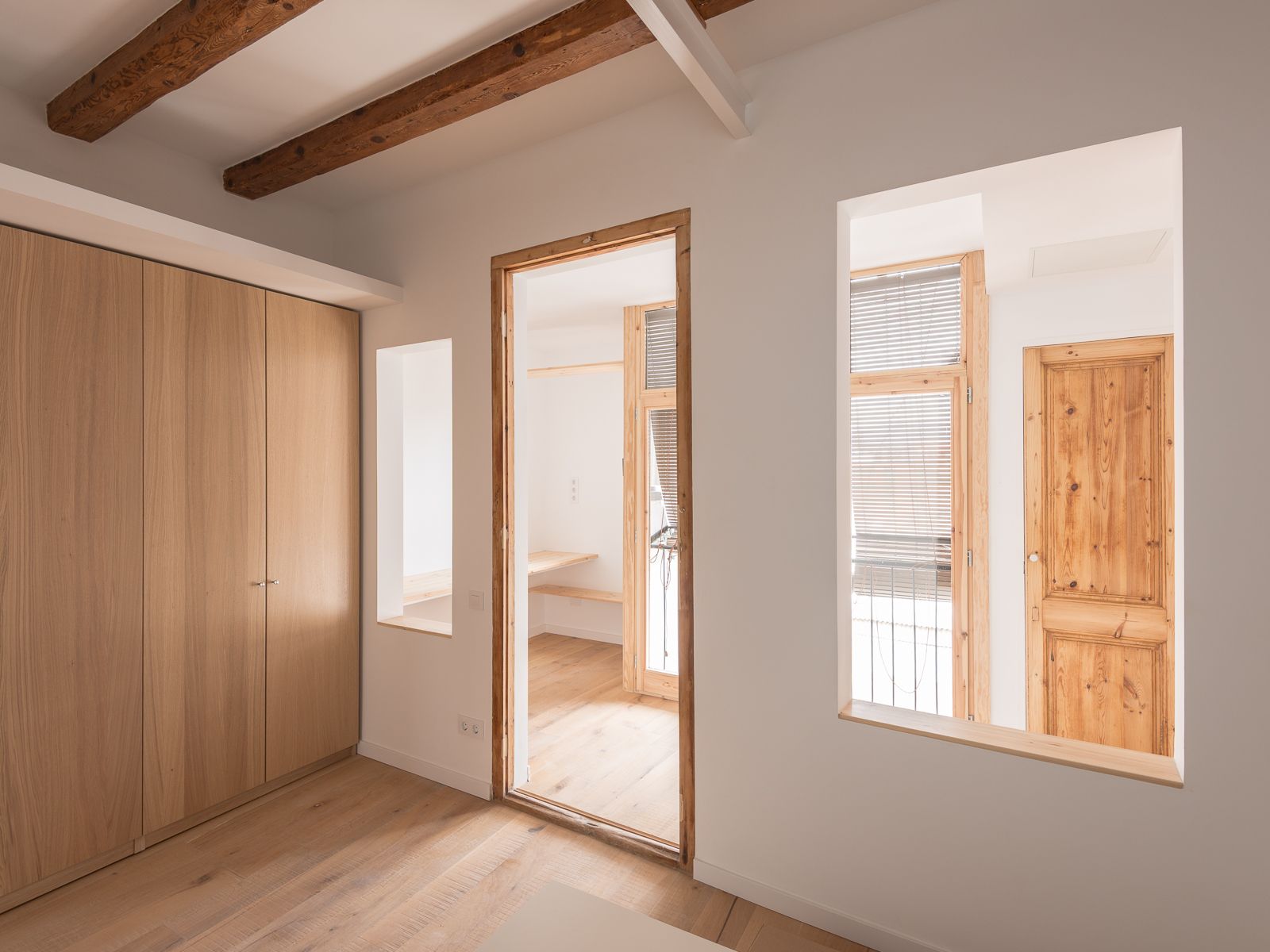 Interior con paredes blancas, vigas de madera expuestas, muebles de madera clara y puertas con vista a una habitación de estudio luminosa con suelos de madera y grandes ventanas.