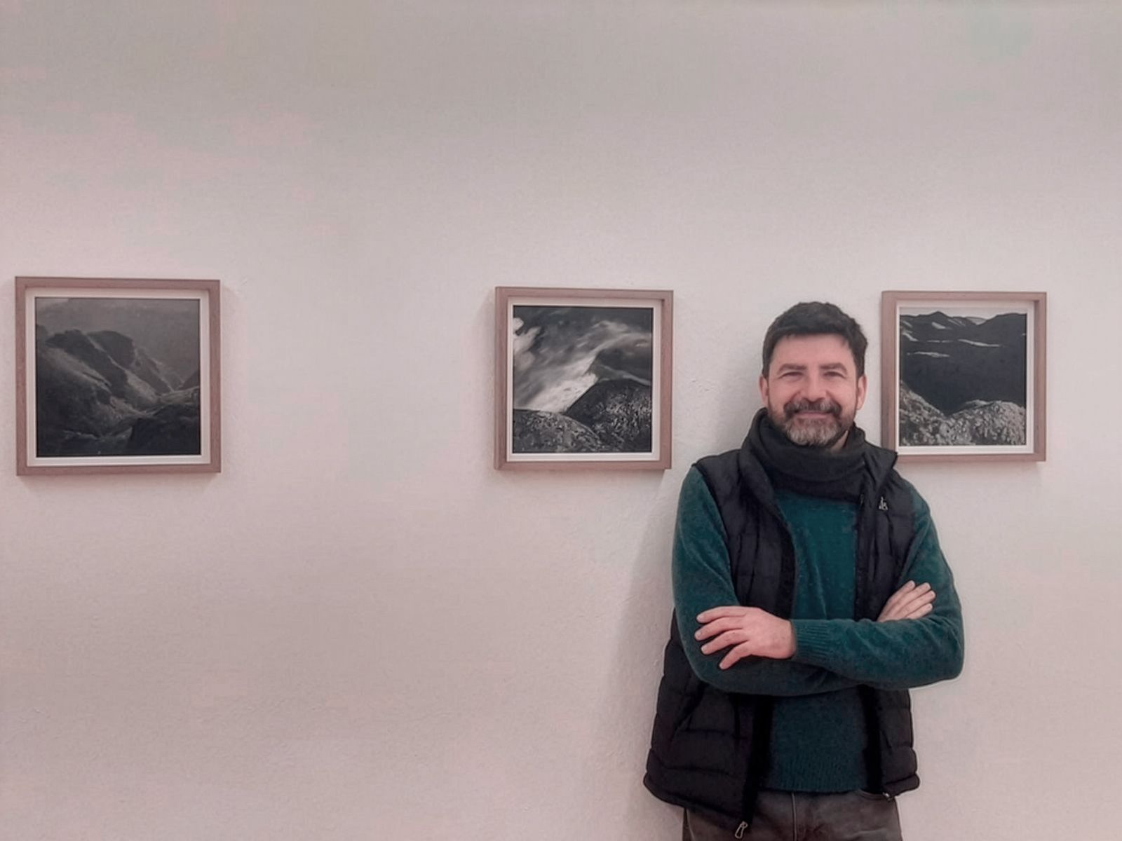 Un home dempeus davant de tres fotografies emmarcades de paisatges, somrient amb els braços creuats.