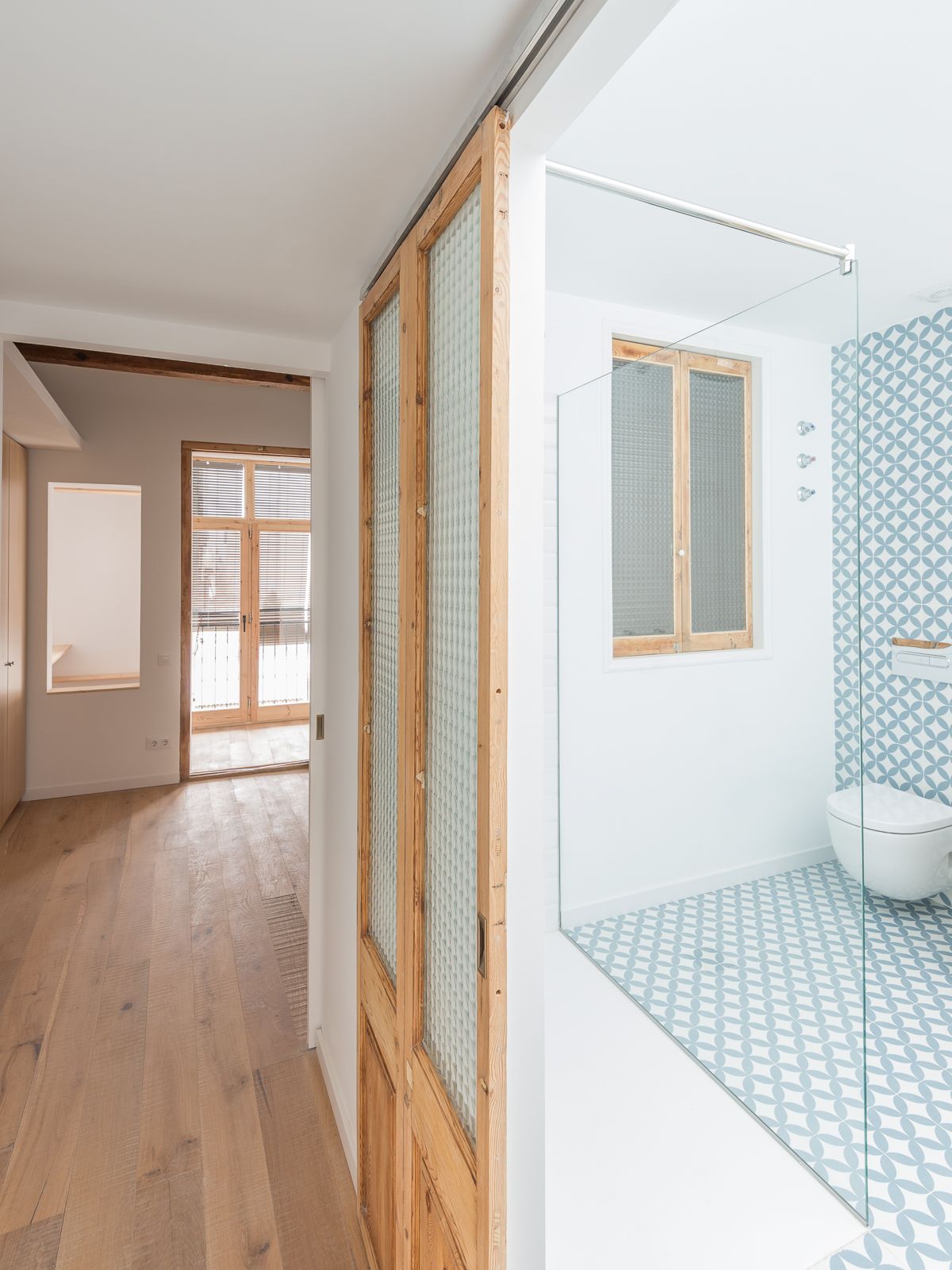 Pasillo con suelos de madera, una puerta corredera de vidrio que conduce a un baño con baldosas con patrones azules y blancos.