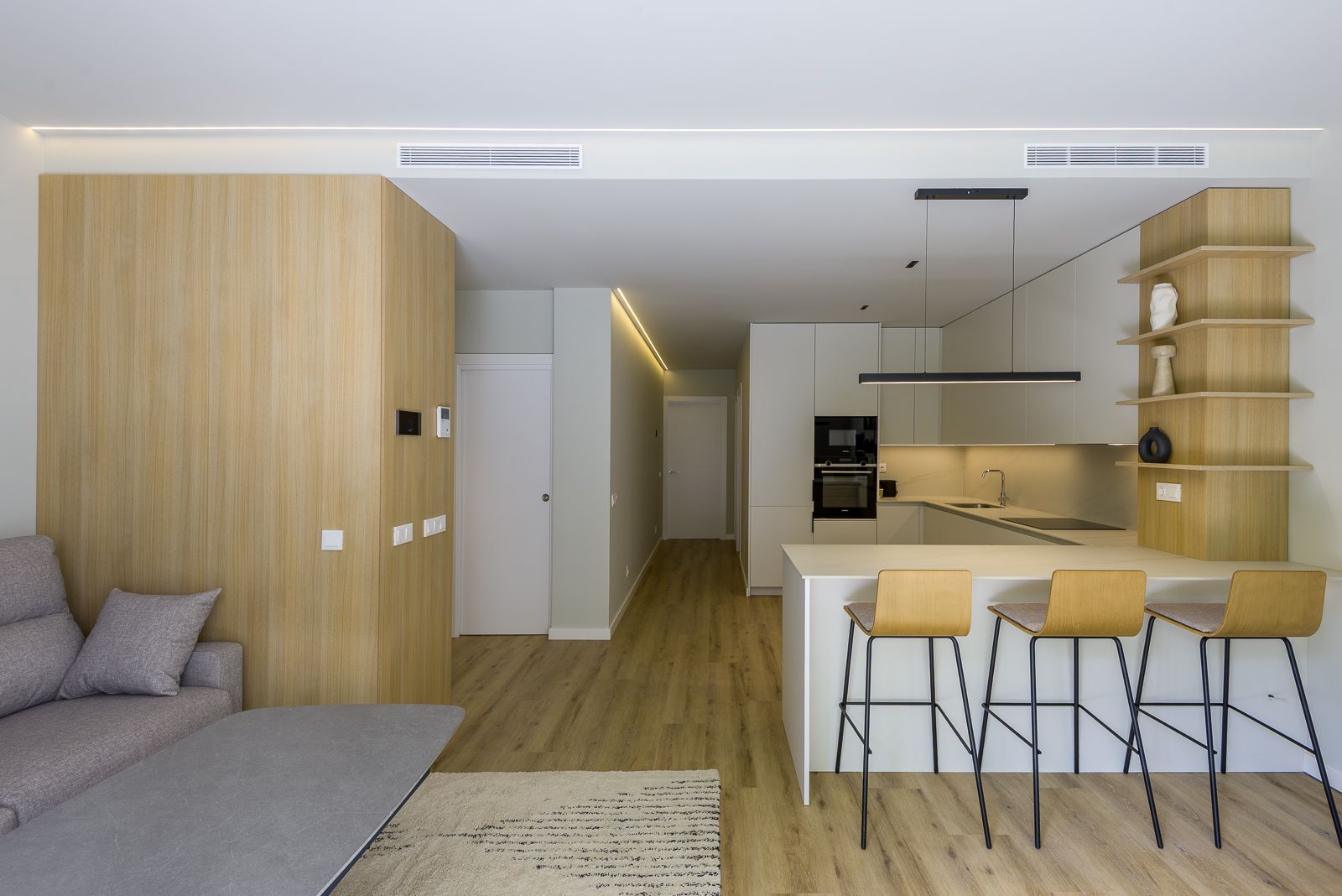 Modern open-concept apartment interior showing a white kitchen peninsula with three light wood barstools, built-in appliances, open shelving, integrated LED ceiling lights, and an adjoining living area with wood-paneled walls and light wood flooring.