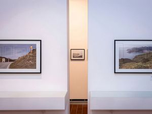 Dos fotografías de paisaje enmarcadas se muestran en una pared blanca con una pequeña fotografía en blanco y negro visible al fondo a través de una abertura.