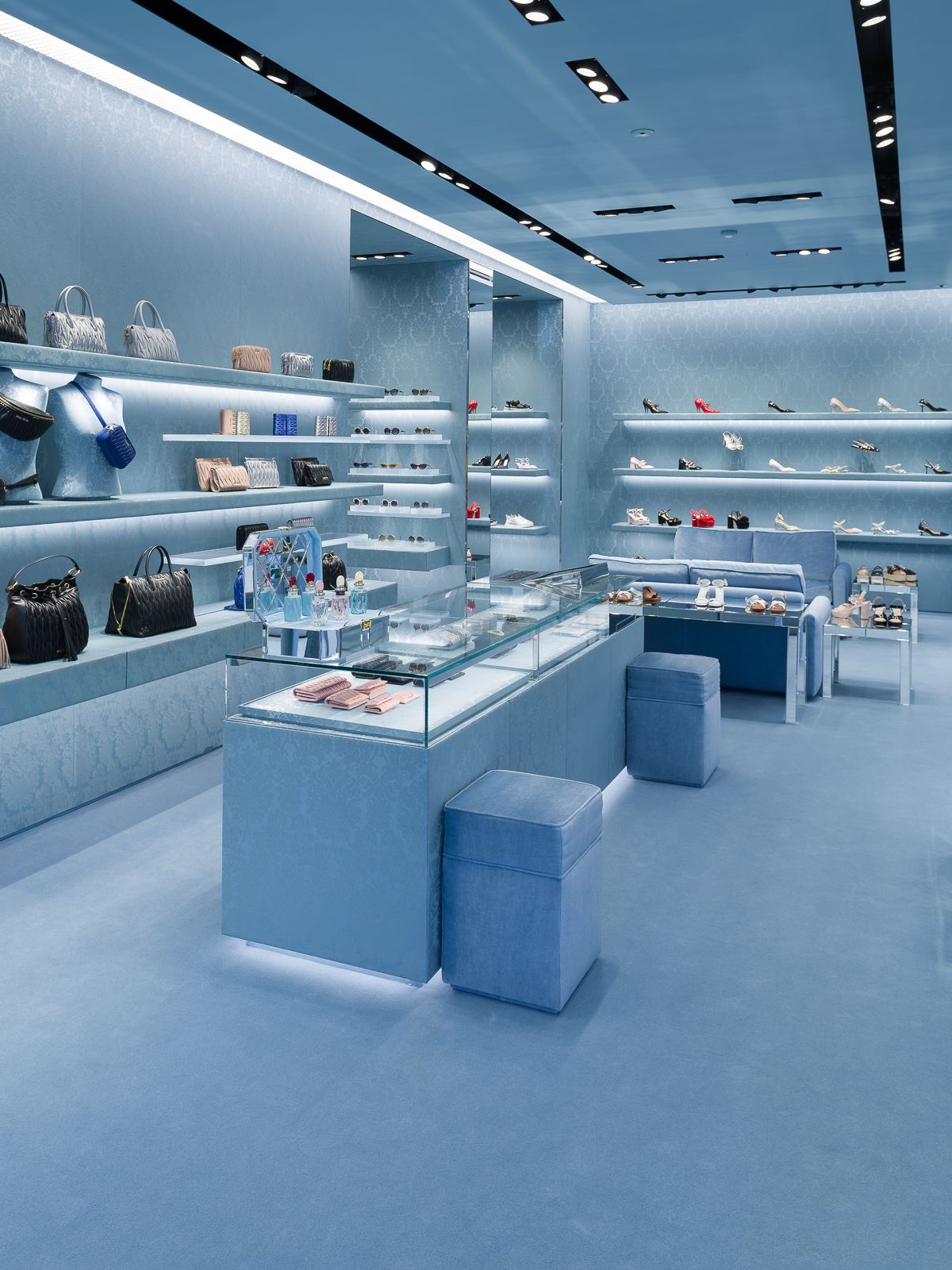 Un interior de tienda Miu Miu elegante, todo en azul, mostrando elegantes estantes de exhibición con bolsos y zapatos, y un mostrador de exhibición de vidrio con accesorios.