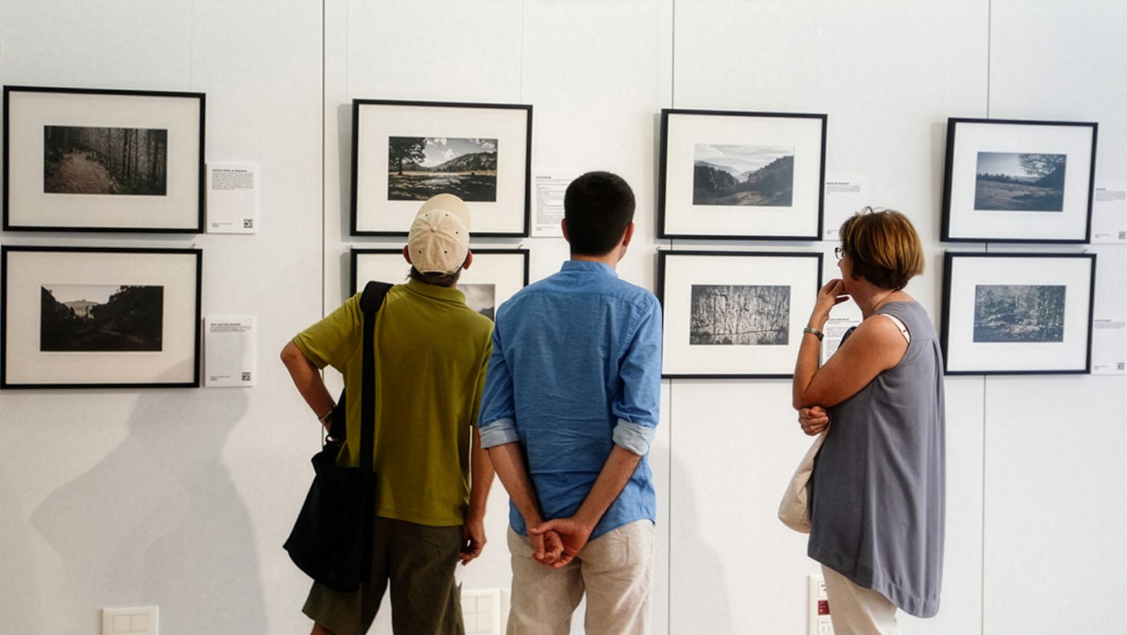 Tres persones examinant de prop fotografies de paisatges emmarcades en una paret de galeria.
