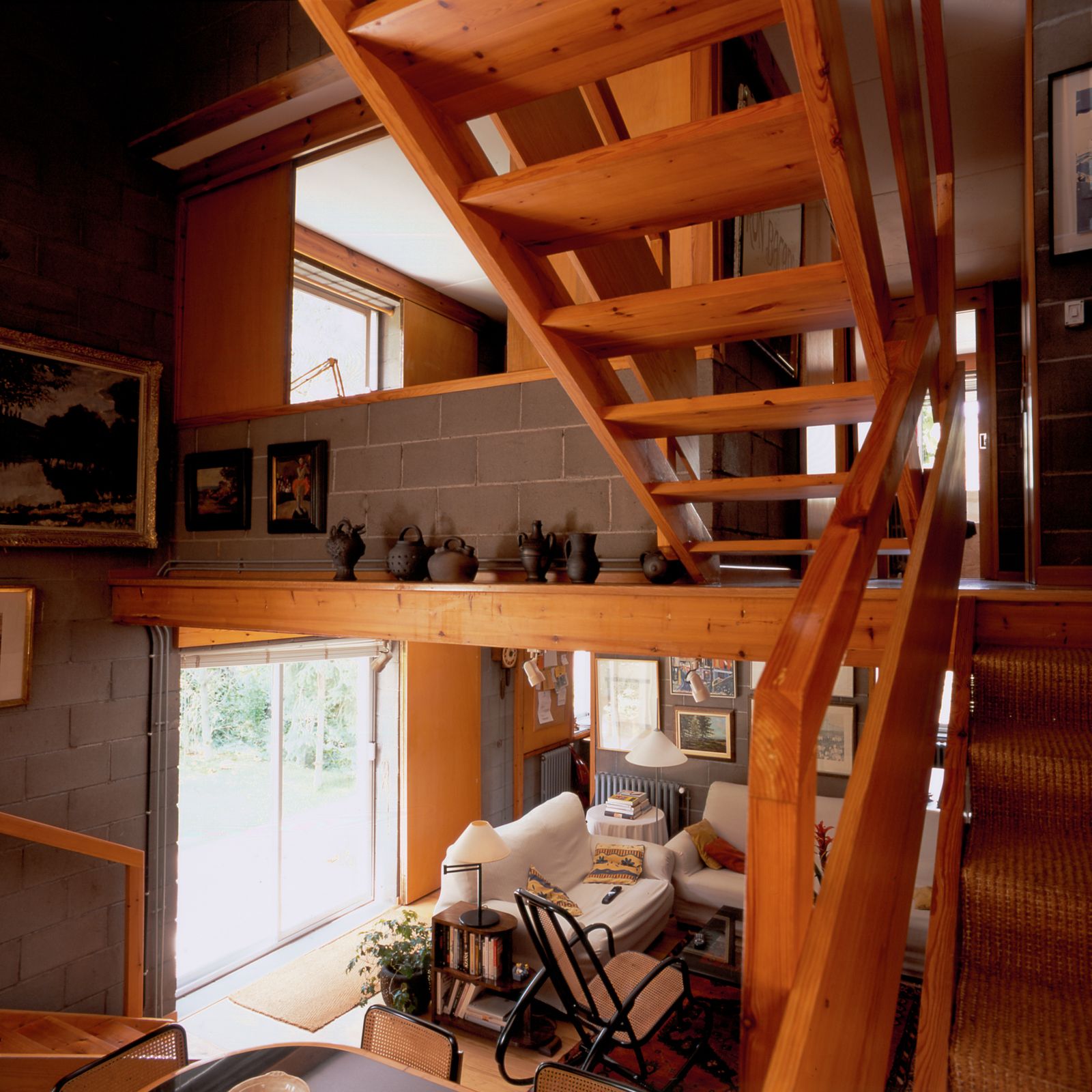 Vista interior que muestra una escalera de madera, una acogedora sala de estar con sofás blancos, una mecedora, una alfombra y objetos decorativos en estantes y paredes. La habitación tiene paredes de bloques de hormigón y una gran puerta corredera que deja entrar luz natural.