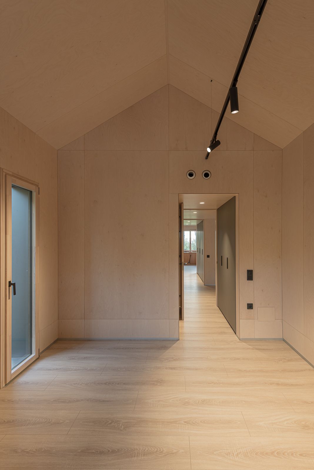 Vista interior de una habitación con techo inclinado, revestimiento de madera clara, iluminación de riel y una puerta que conduce a un pasillo, con una ventana en el extremo.