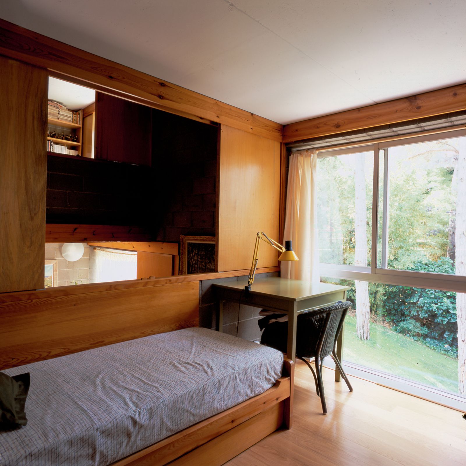 Dormitorio con una cama individual de madera, paredes de madera, escritorio verde oscuro con una lámpara amarilla, gran ventana de suelo a techo con vistas a un jardín y un panel corredero de pared abierto con vistas a un espacio central.