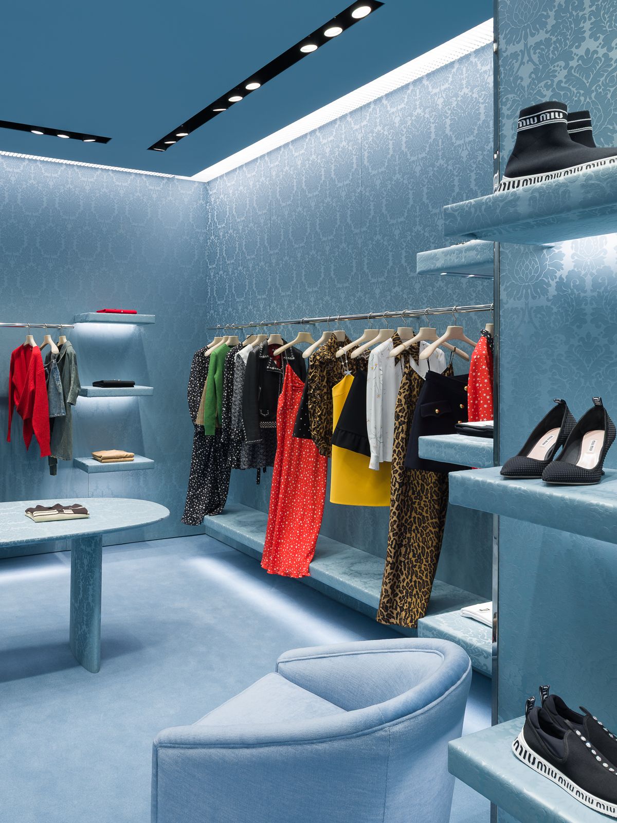 Un lujoso interior de tienda Miu Miu con el color azul en paredes estampadas, sofás y mesas, así como suelo y techo. Una variedad de ropa colorida exhibida en percheros y zapatos elegantes exhibidos en elegantes estantes.