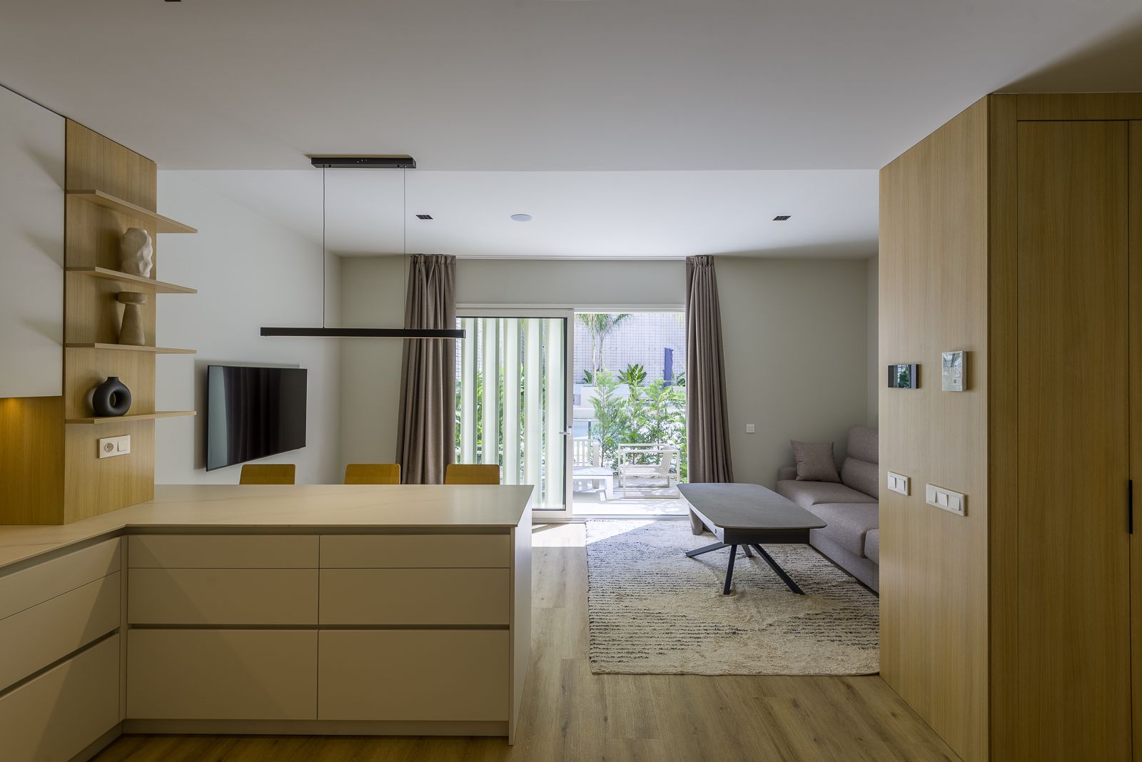 Bright, open-concept living space viewed over a beige kitchen peninsula, showing floating light wood shelves, a sleek black linear pendant light, a grey couch with a low coffee table, and sliding glass doors opening to a sunlit garden patio.