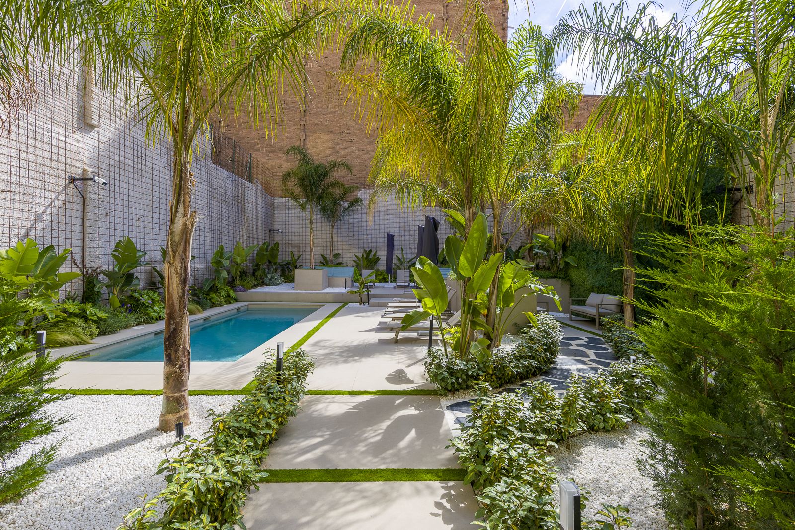 Lush tropical courtyard patio featuring a paved walkway lined with low hedges, tall palm trees, and white gravel, leading toward an outdoor swimming pool area with sun loungers and high brick boundary walls covered in climbing wire trellises.