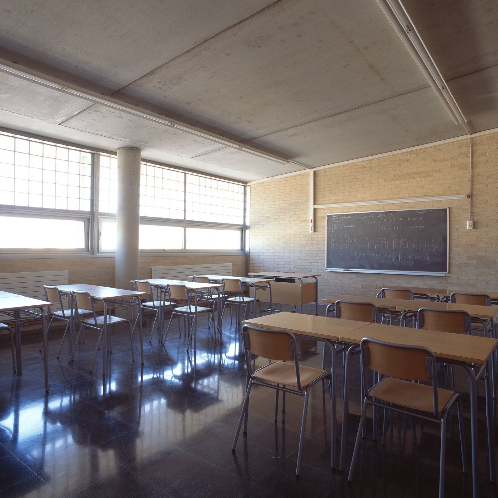 Classroom with wooden desks and chairs, a large blackboard, and windows allowing natural light to illuminate the space.