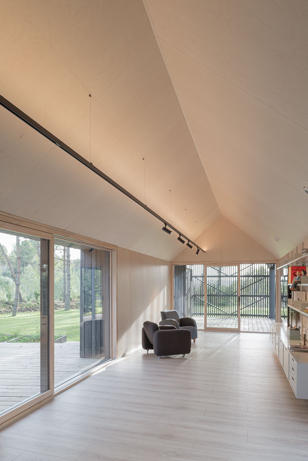 Un área de estar moderna con un techo alto inclinado, iluminación de riel, suelo de madera clara y grandes puertas y ventanas de vidrio, con dos sillones grises.