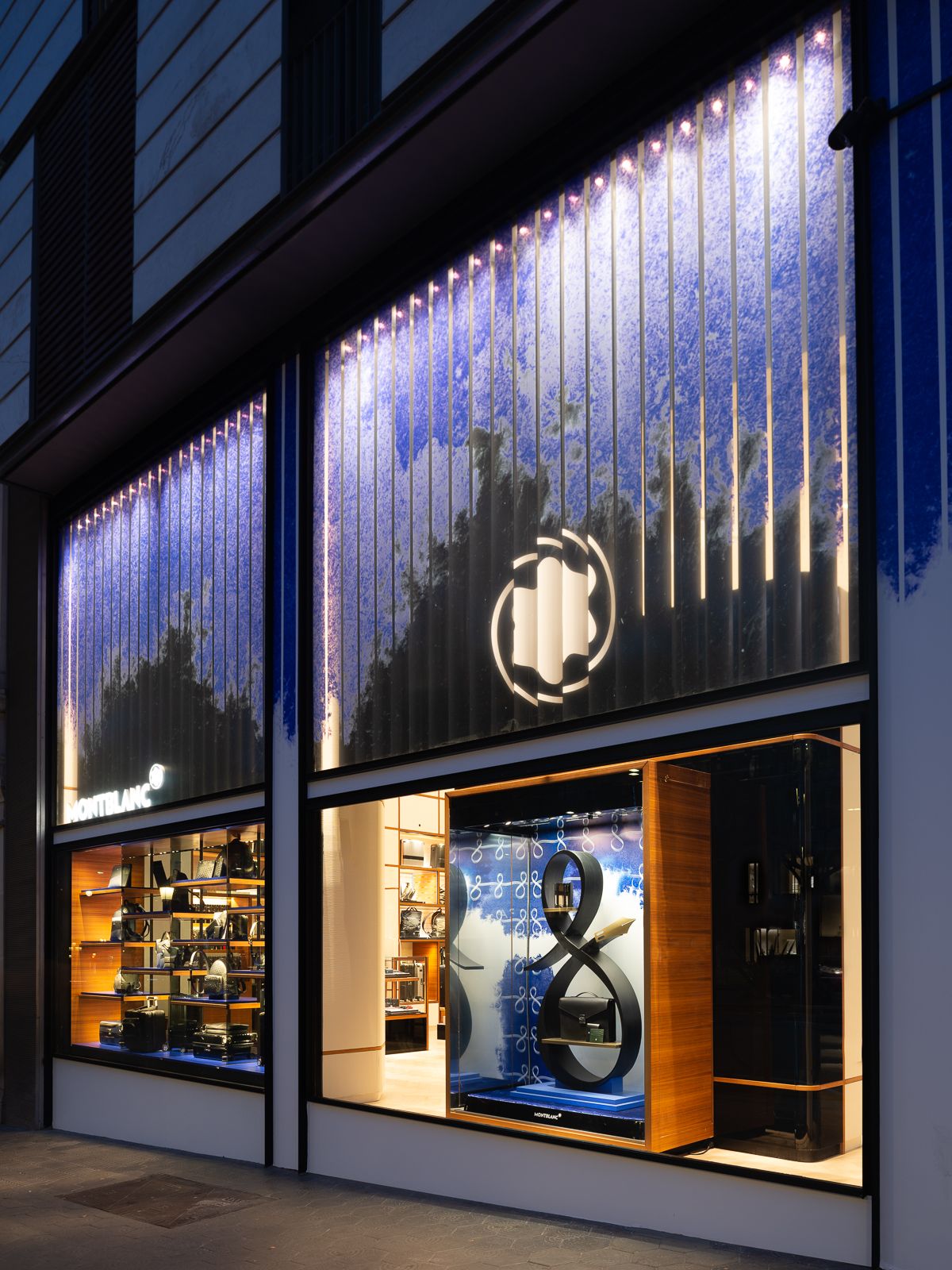Exterior de tienda Montblanc con escaparate iluminado con una escultura que se asemeja a un símbolo de infinito vertical y productos sobre fondo azul.
