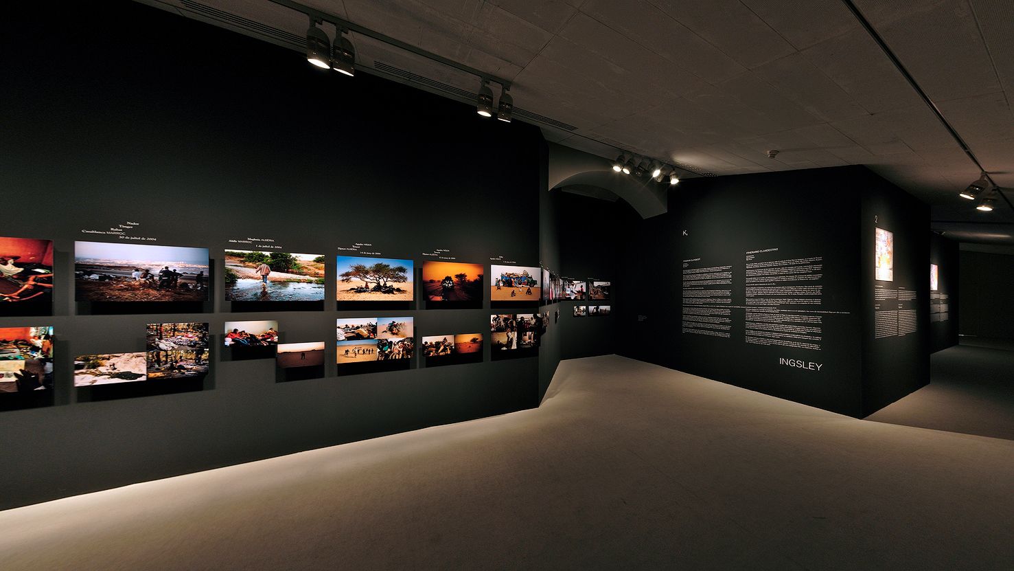 Una galería que muestra una serie de fotografías y paneles de texto descriptivo en paredes oscuras, iluminados por focos.