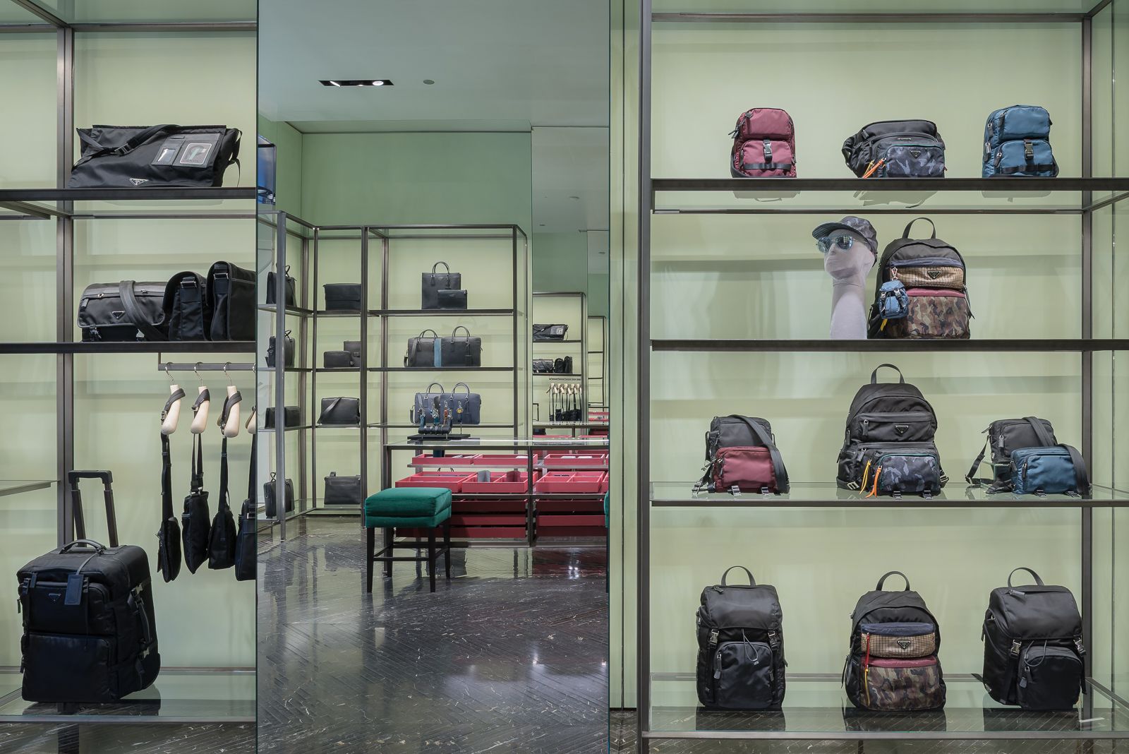 Interior de tienda de lujo con espejos mostrando varios bolsos y mochilas de diseño exhibidos en elegantes estantes metálicos contra un fondo verde pálido.