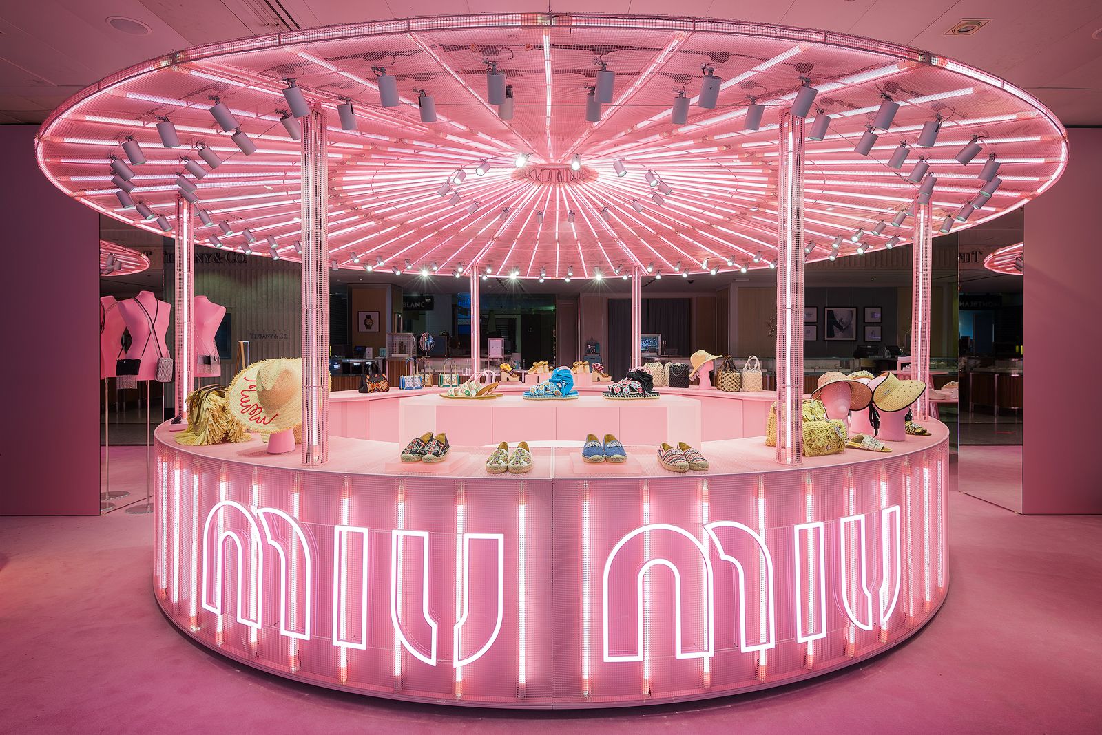 Exhibición iluminada de la tienda pop-up rosa de Miu Miu con bolsos, zapatos y sombreros bajo un dosel de luces rosas brillantes.