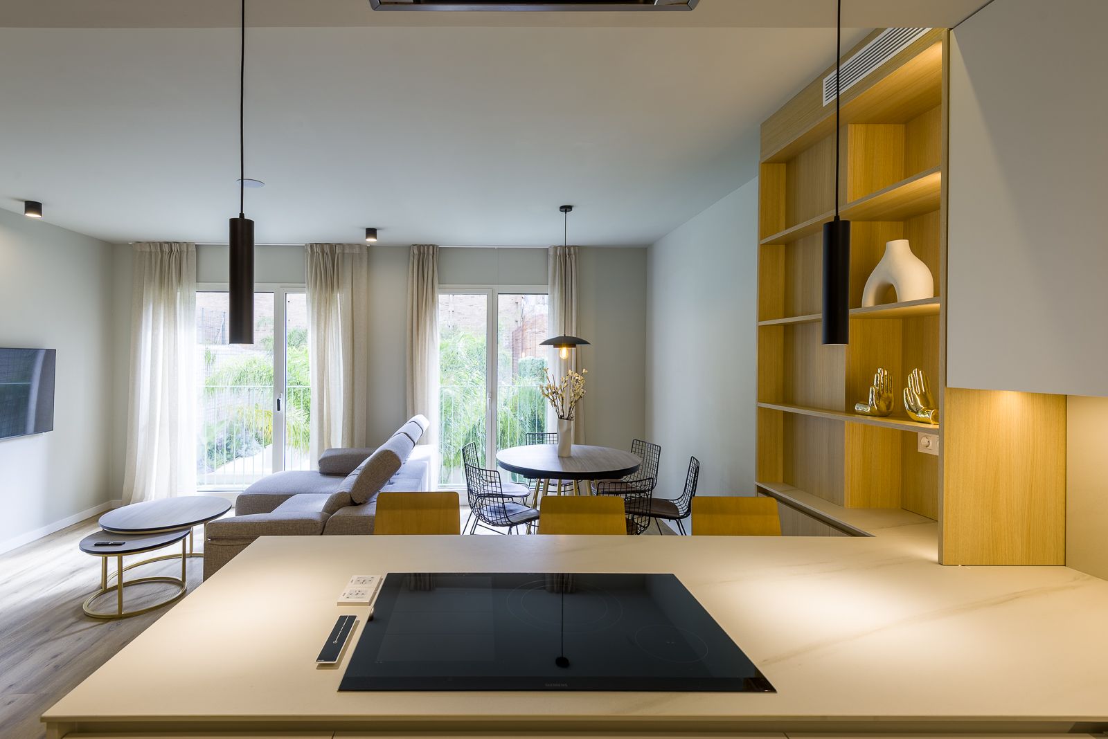 View from a white kitchen island with a built-in cooktop, looking into a modern open-plan living and dining space with light wood shelving, a grey sectional sofa, and floor-to-ceiling glass doors opening to greenery.
