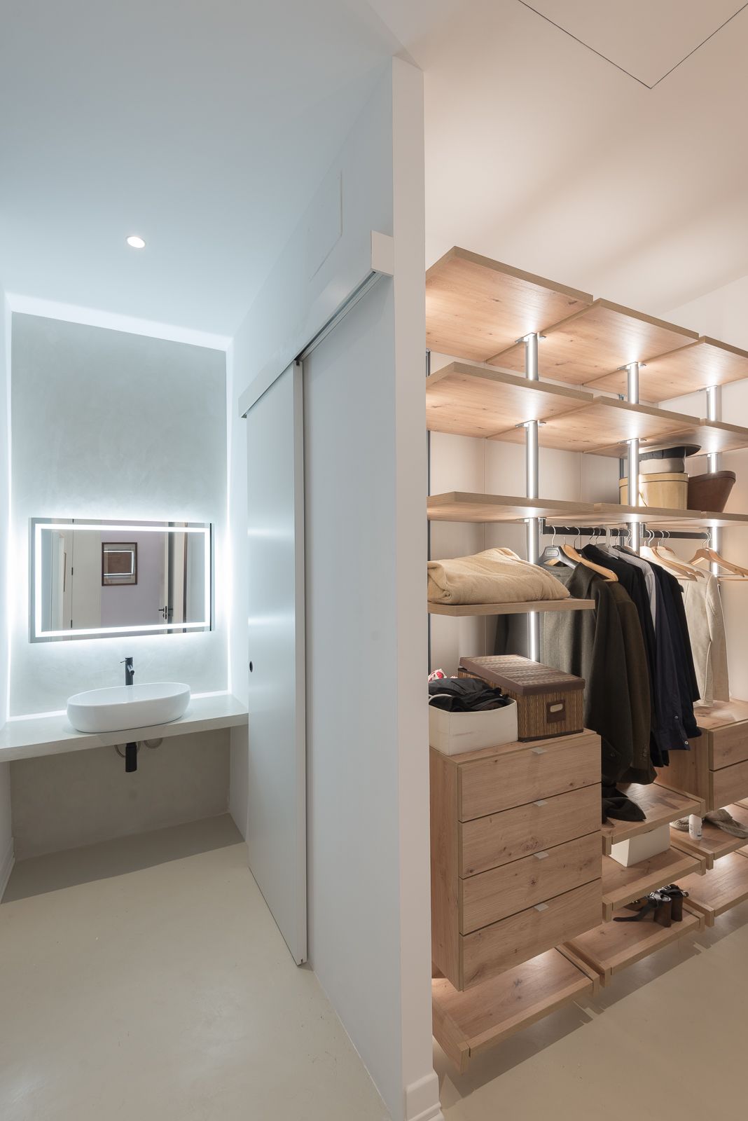 Walk-in closet with wooden shelves and drawers, adjacent to a bathroom with a backlit mirror.