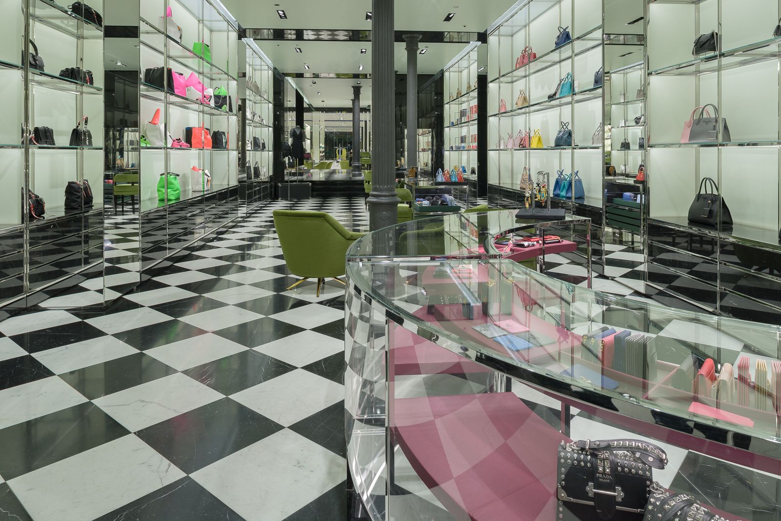 Interior de tienda Prada con suelo de tablero de ajedrez blanco y negro, estantes de vidrio que exhiben bolsos coloridos, sillas verdes y columnas de hierro fundido negro.