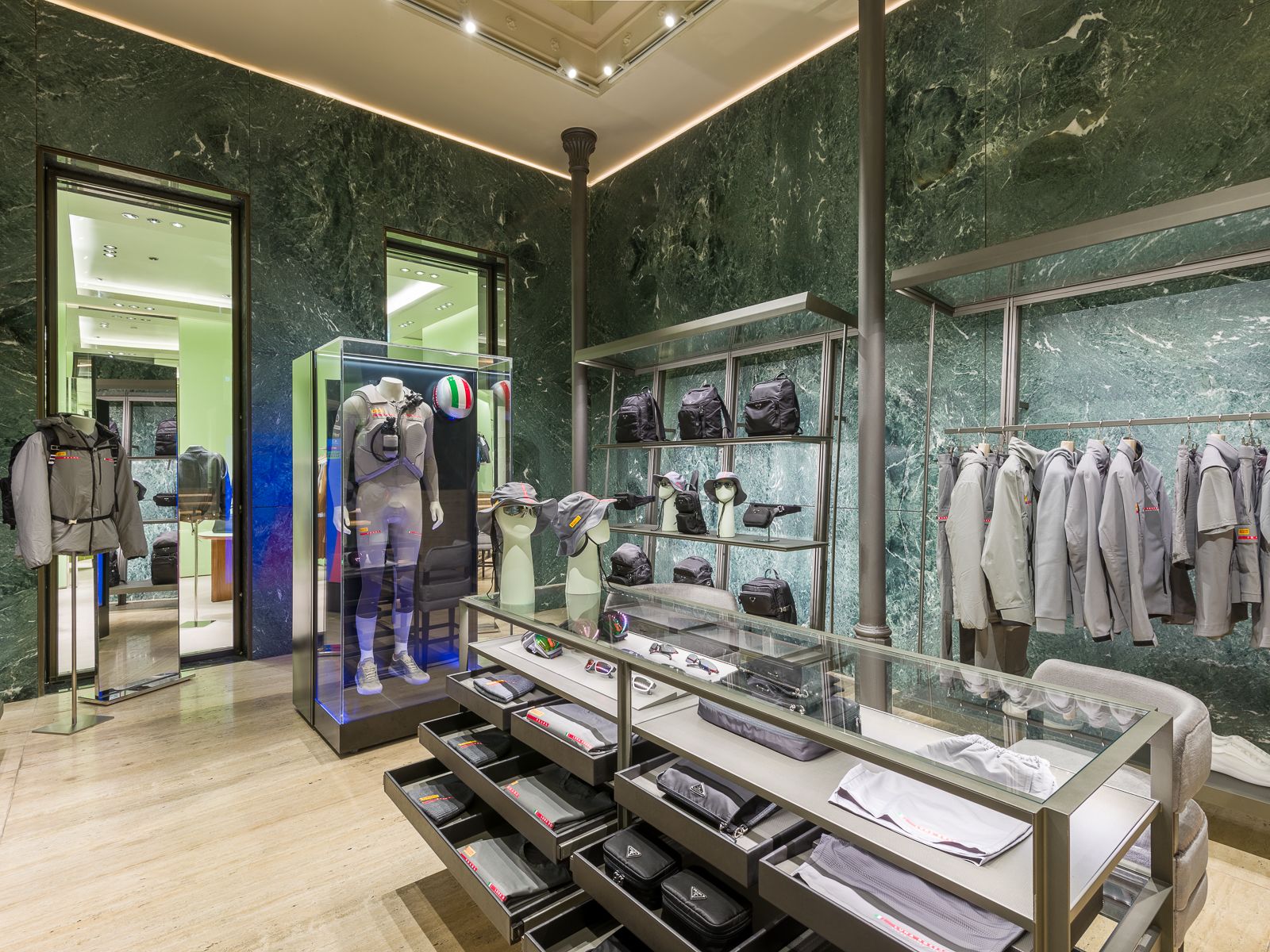 Lujosa boutique Prada con paredes de mármol verde, mostrando zapatillas de diseño, bolsos negros y accesorios en elegantes estanterías metálicas. Una mesa de vidrio exhibe gafas y sombreros, mientras que un maniquí en un atuendo de rendimiento está encerrado en una vitrina de vidrio.