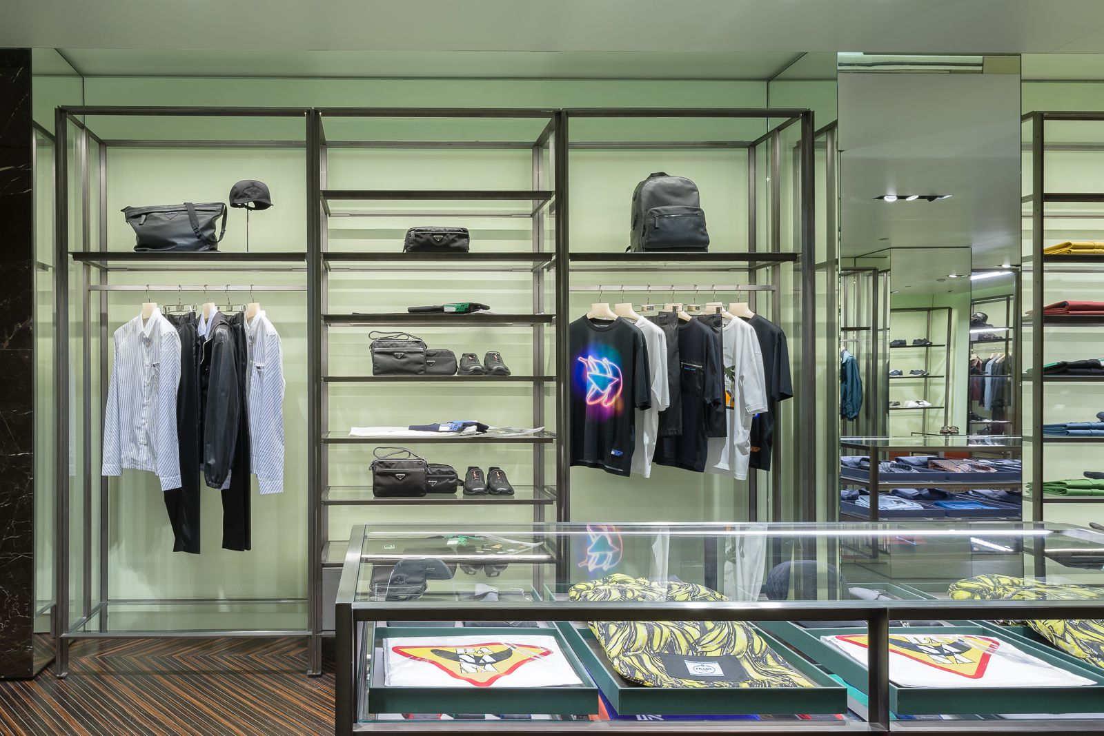 Interior de tienda minorista de lujo con estantes ordenadamente organizados que exhiben bolsos de diseño, ropa y accesorios contra un fondo verde Prada.