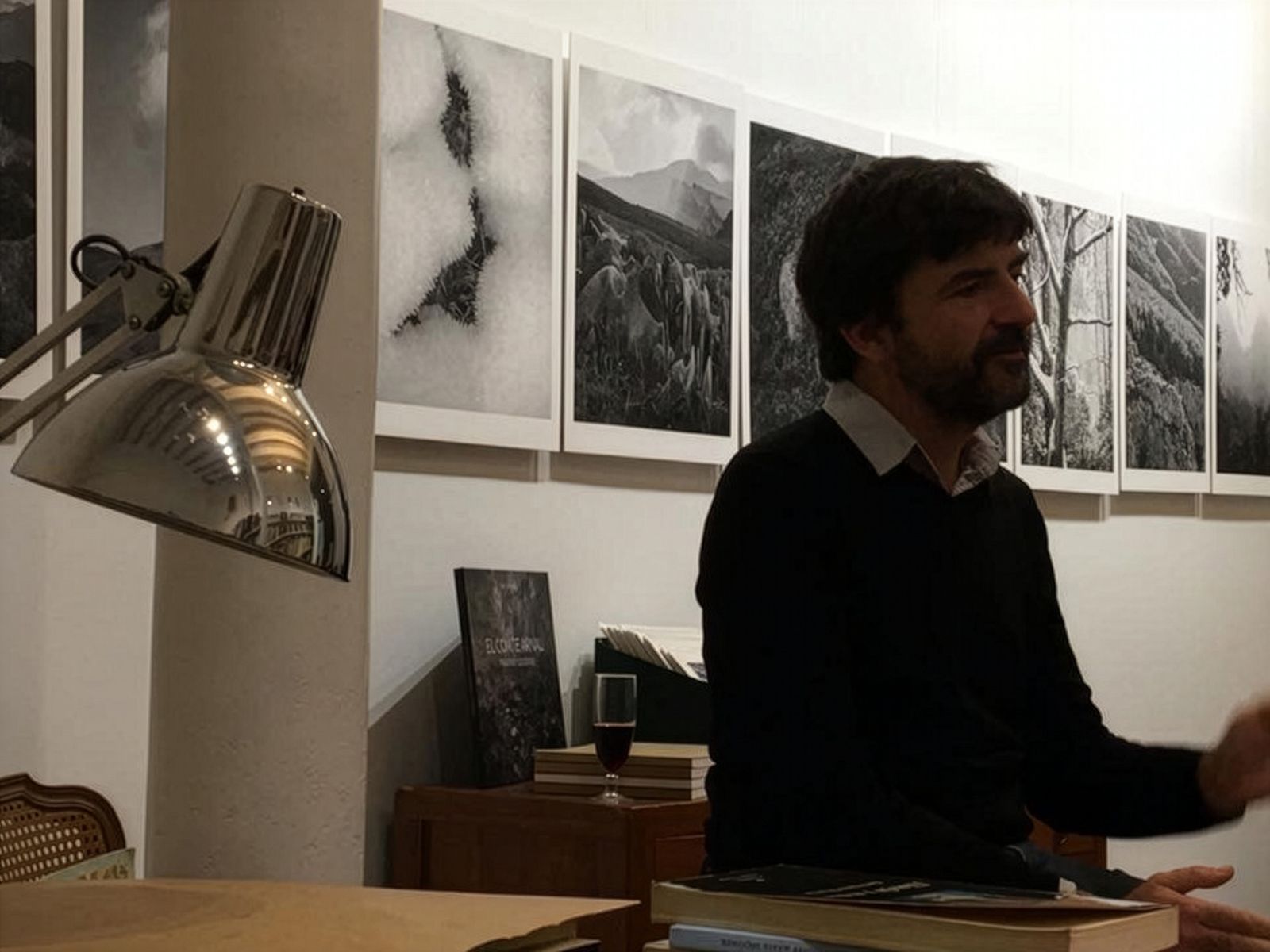 Un hombre sentado frente a una exposición de fotografías en blanco y negro de paisajes montañosos en un acogedor espacio de exposición. Una lámpara de escritorio está colocada cerca, y una copa de vino reposa sobre un mueble detrás de él.