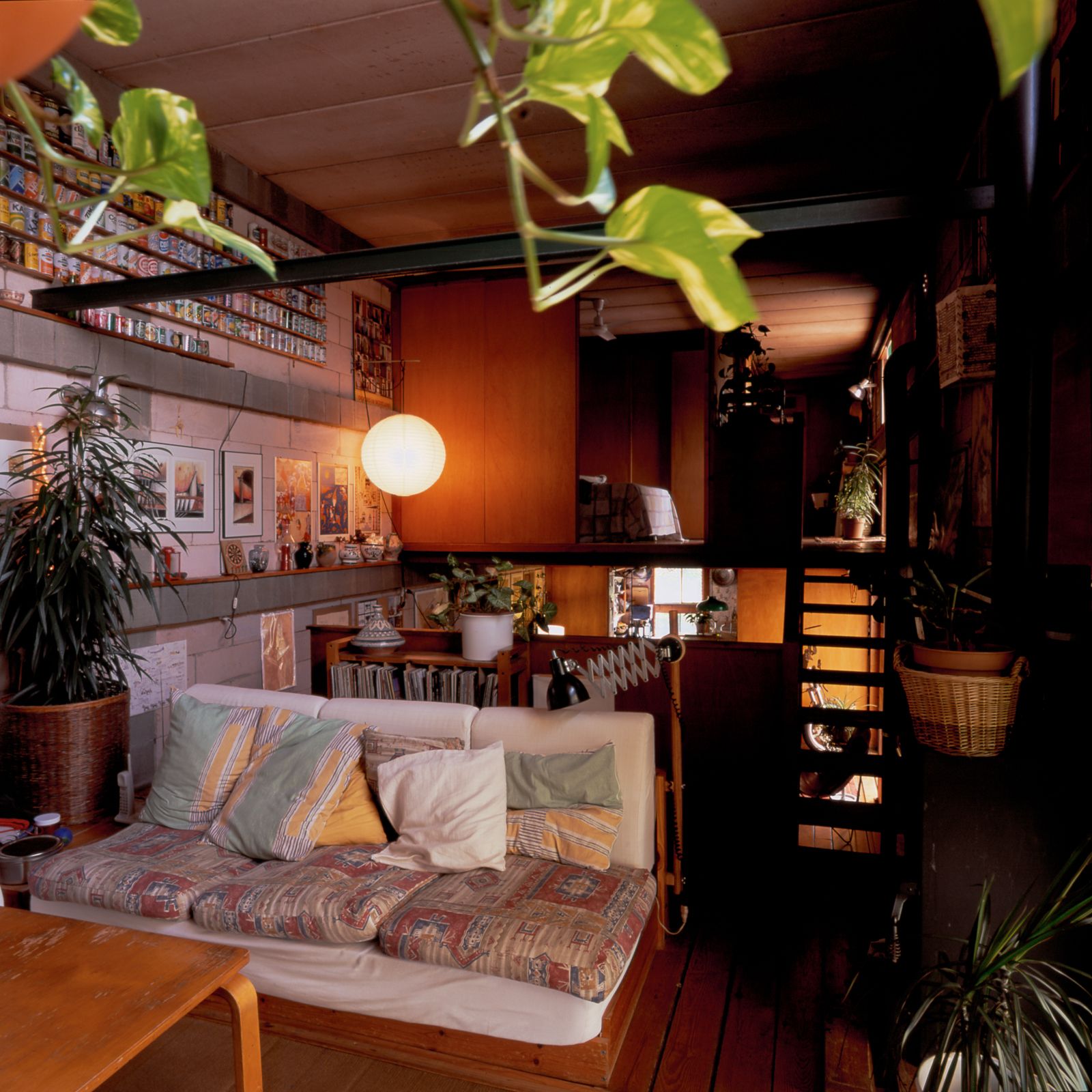 Sala de estar acogedora con un sofá colorido, muebles de madera, plantas de interior y una lámpara colgante blanca, con una escalera y una pared decorada con arte y coleccionables.