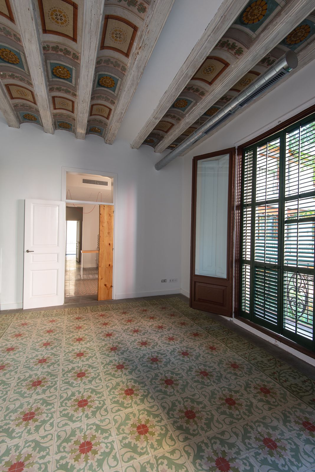 Habitación con vigas de techo ornamentadas y baldosas de suelo estampadas, con una puerta de madera parcialmente abierta y una ventana con contraventanas.