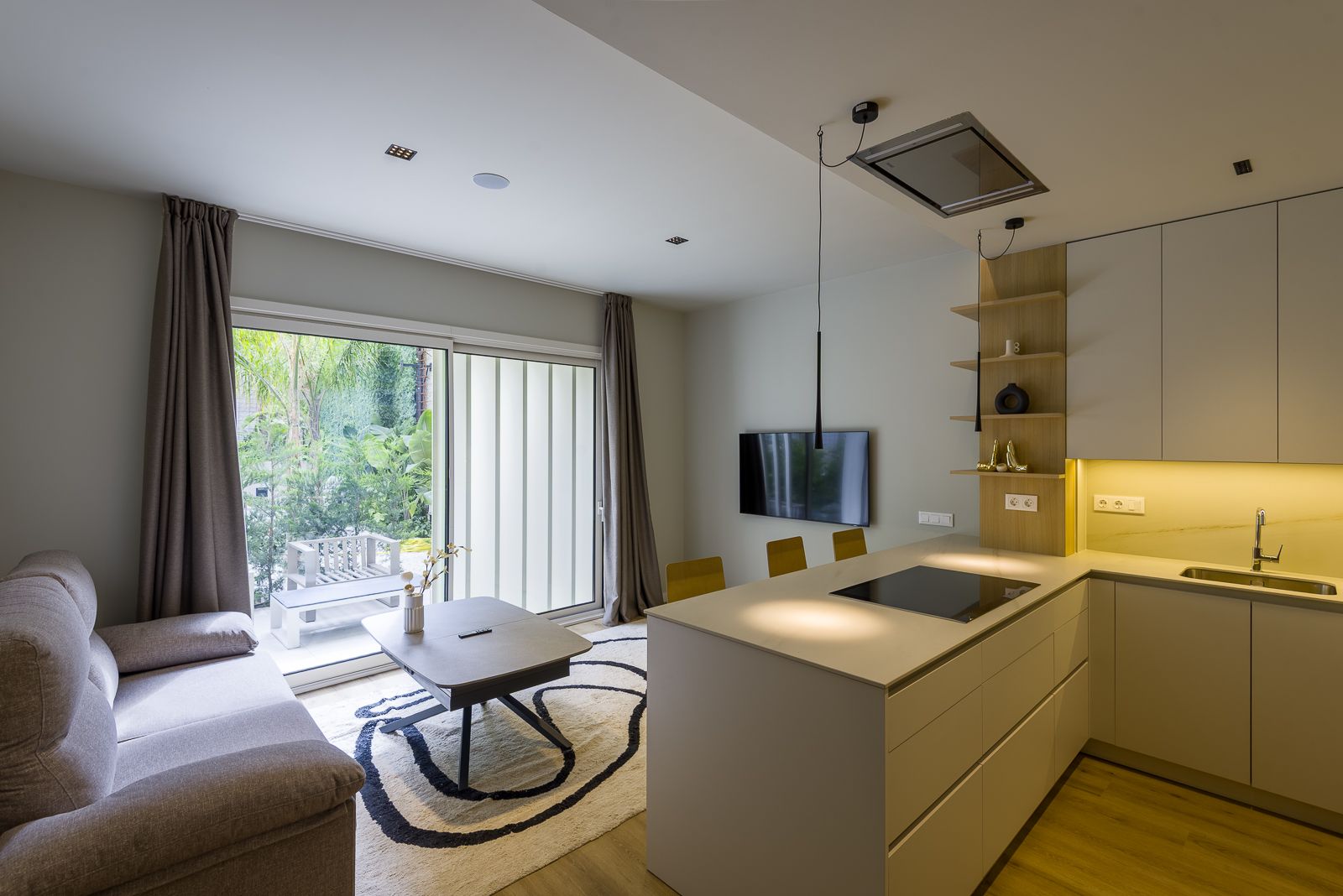 Sleek open-concept interior showing a modern cream-colored kitchen with an island peninsular cooktop and under-cabinet lighting, adjacent to a living room with a plush grey sofa, low coffee table, and sliding glass doors revealing a lush green patio.