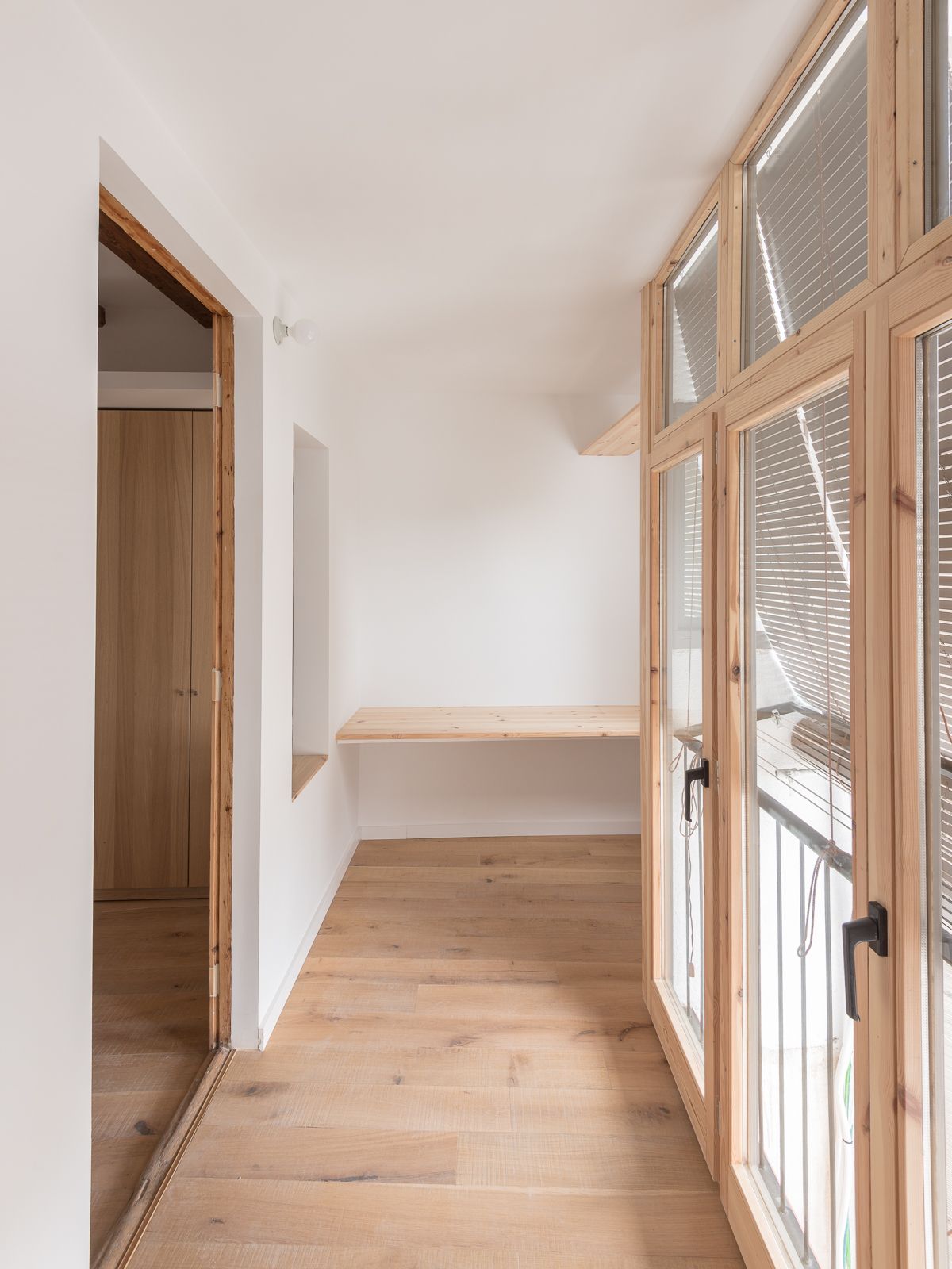 Pequeña área de estudio con suelos de madera, un escritorio de madera integrado y grandes ventanas de balcón de suelo a techo con persianas.
