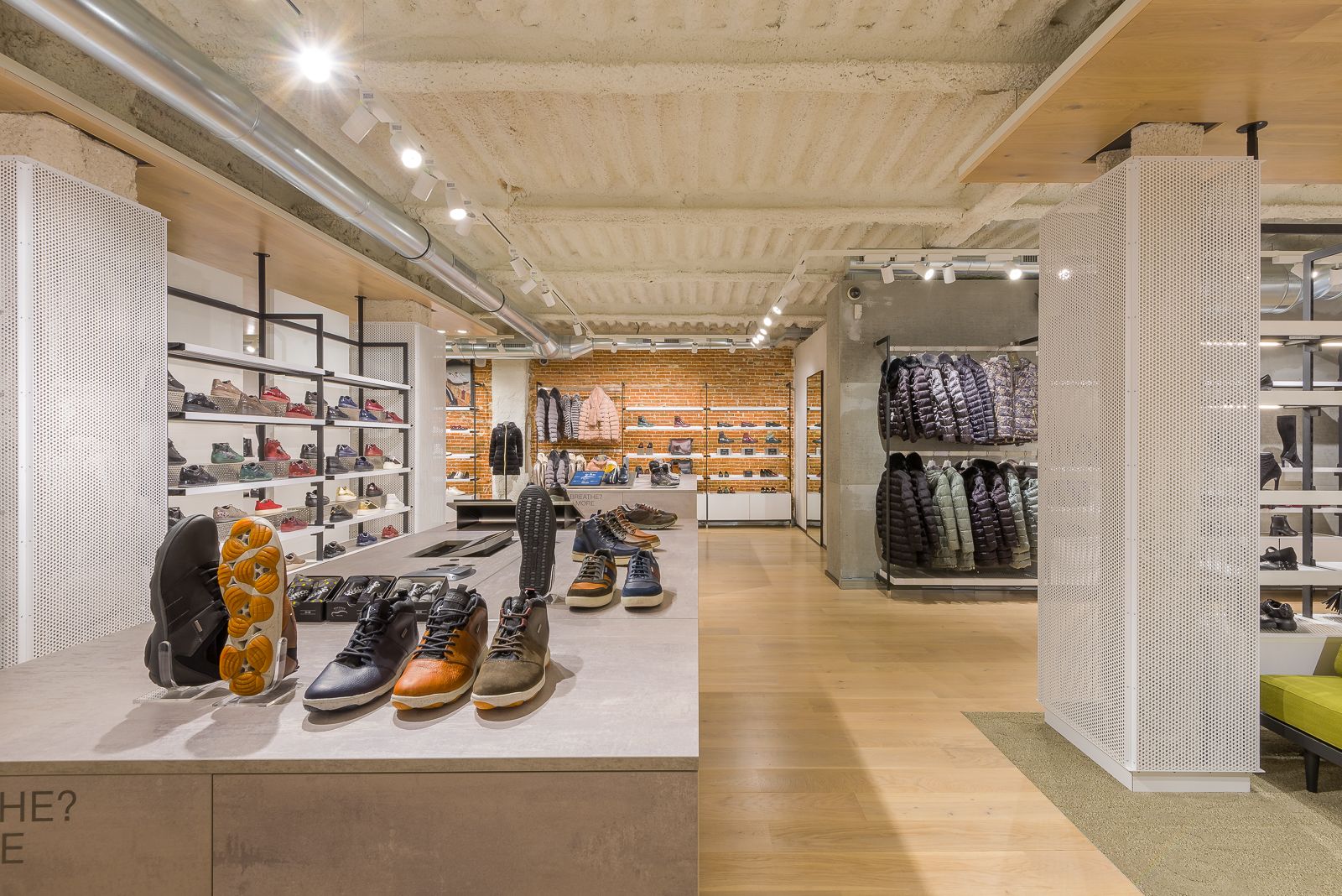 Interior de la tienda Geox mostrando un surtido de zapatos en mesas de exhibición, estantes con zapatos y percheros de chaquetas, con un techo industrial moderno y pared de ladrillo expuesto al fondo.