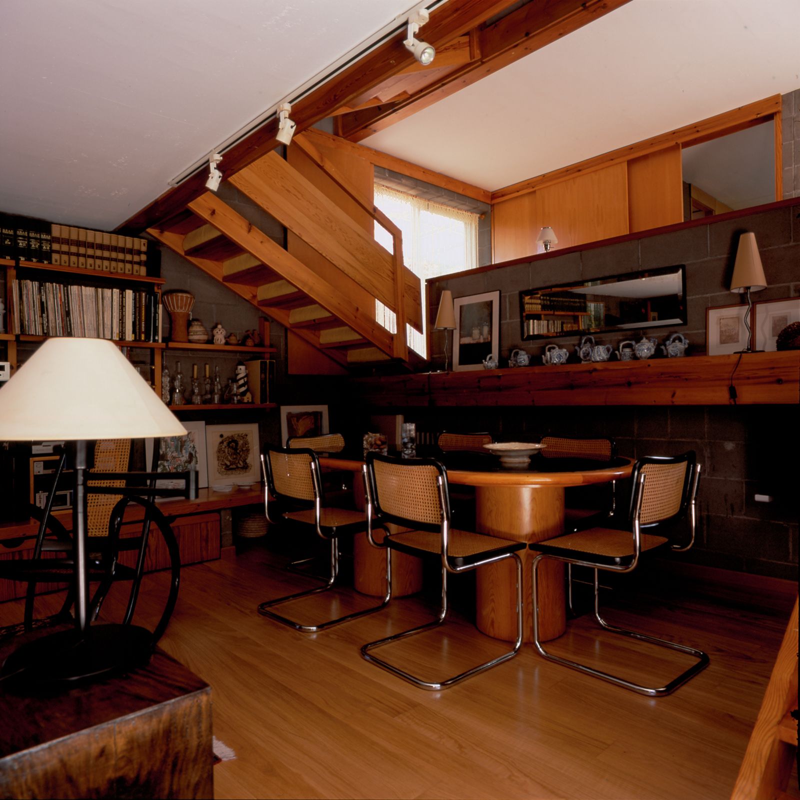 Comedor con mesa de madera ovalada, seis sillas Cesca, escalera de madera que conduce al piso superior, estanterías y varios objetos decorativos en los estantes.