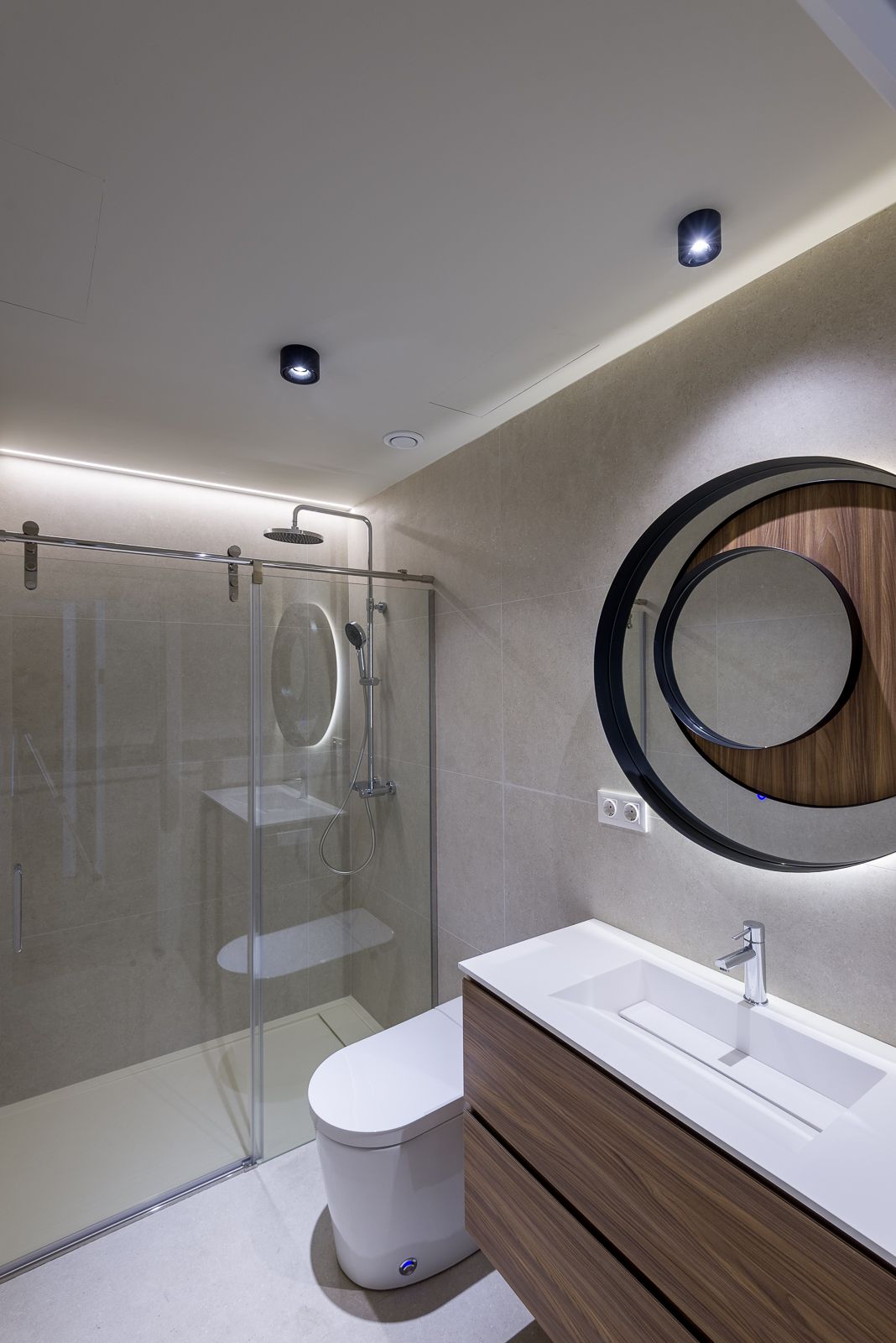 Contemporary bathroom with beige tiled walls, featuring a wooden vanity cabinet with a white integrated sink, a large backlit circular mirror, a modern white toilet, and a glass-enclosed shower stall with a rain shower head.