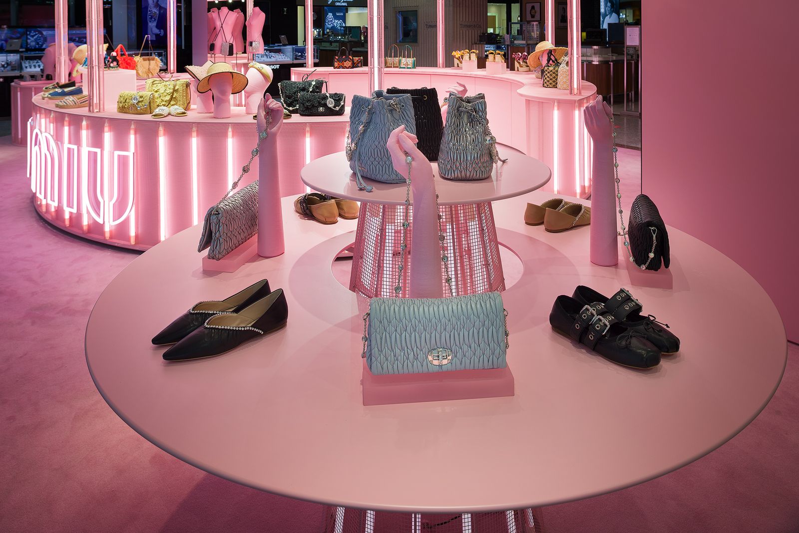 Primer plano de una exhibición de tienda pop-up de Miu Miu de temática rosa, con bolsos y zapatos dispuestos en una mesa redonda de dos niveles con una base cónica iluminada desde dentro por iluminación de neón. Sobre la mesa, tres manos de maniquí rosas mostrando accesorios.