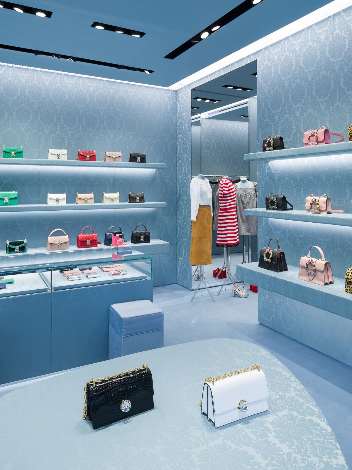 Un elegante interior de tienda Miu Miu, con elegantes estantes de exhibición con una variedad de bolsos coloridos, mostradores de vidrio que exhiben accesorios más pequeños y, en primer plano, un bolso negro junto a un bolso blanco en una mesa ovalada con patrón azul.
