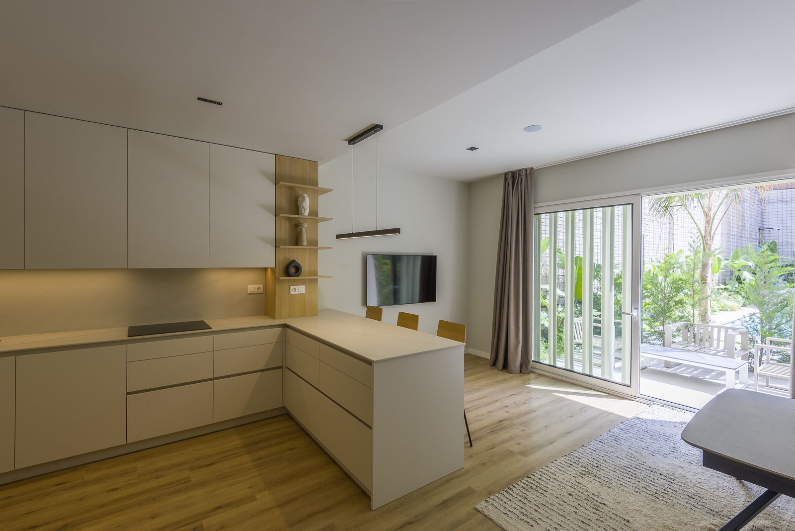Modern open-concept interior featuring full-height beige kitchen cabinets, an L-shaped counter with integrated cooktop and breakfast bar, a wall-mounted TV, and large glass sliding doors with vertical louvers opening to a sunlit garden patio with palm trees.