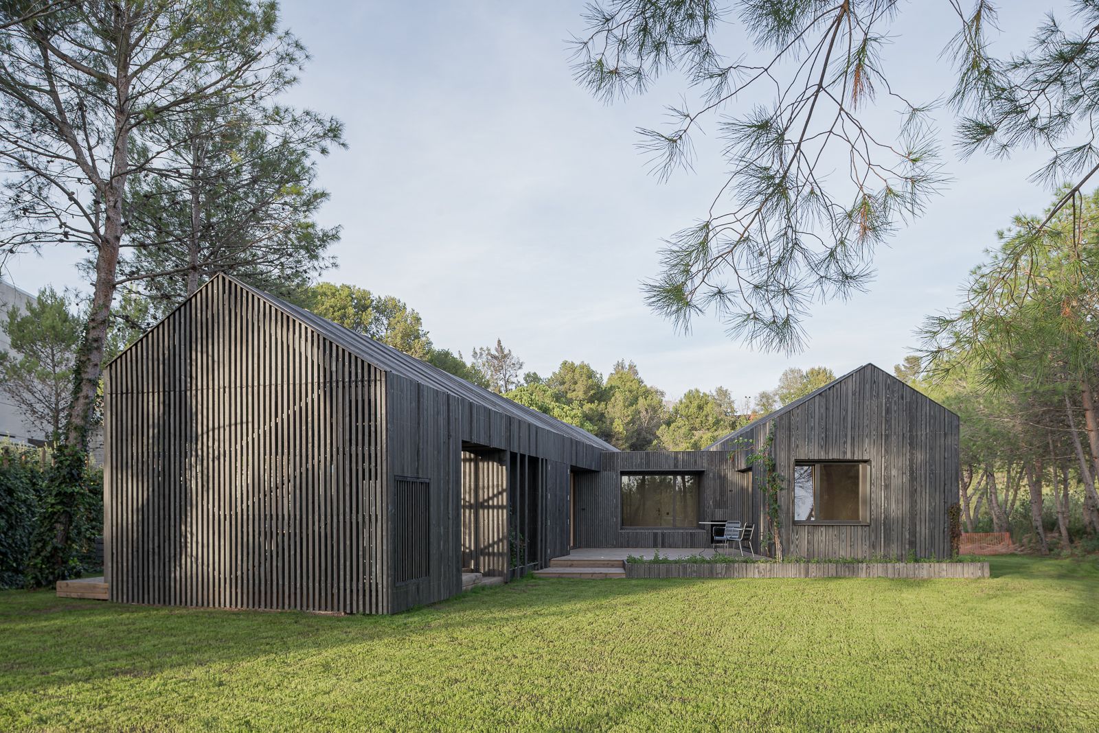 Vista exterior de una casa moderna con dos cuerpos, techo inclinado, revestimiento de madera oscura, grandes ventanas y un amplio césped rodeado de pinos.