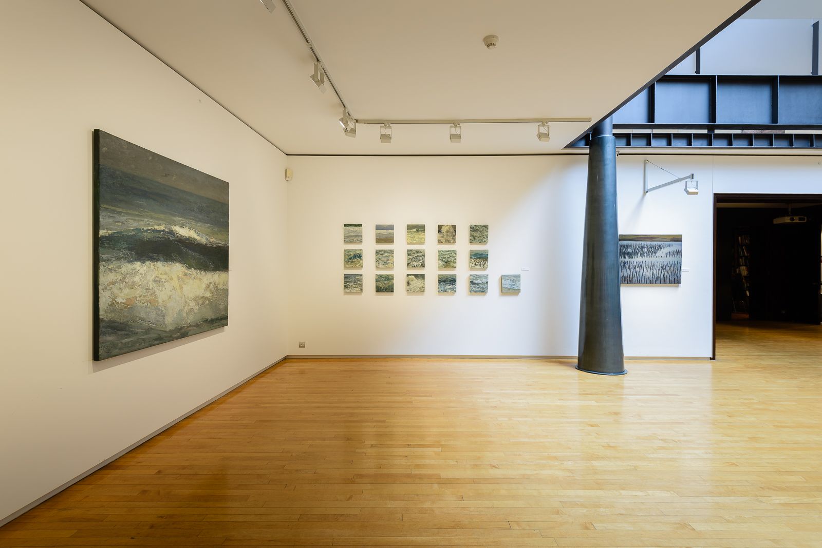 Una espaciosa galería de arte con paredes blancas y suelos de madera pulida, exhibiendo pinturas de paisajes marinos bajo iluminación de riel. Una columna de acero cónica negra se encuentra al lado.
