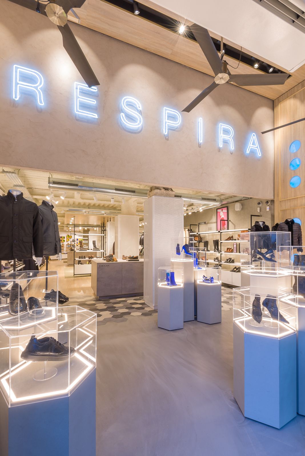 Vista interior de la tienda Geox, con exhibiciones de zapatos iluminadas y un gran letrero de neón 'RESPIRA' arriba.