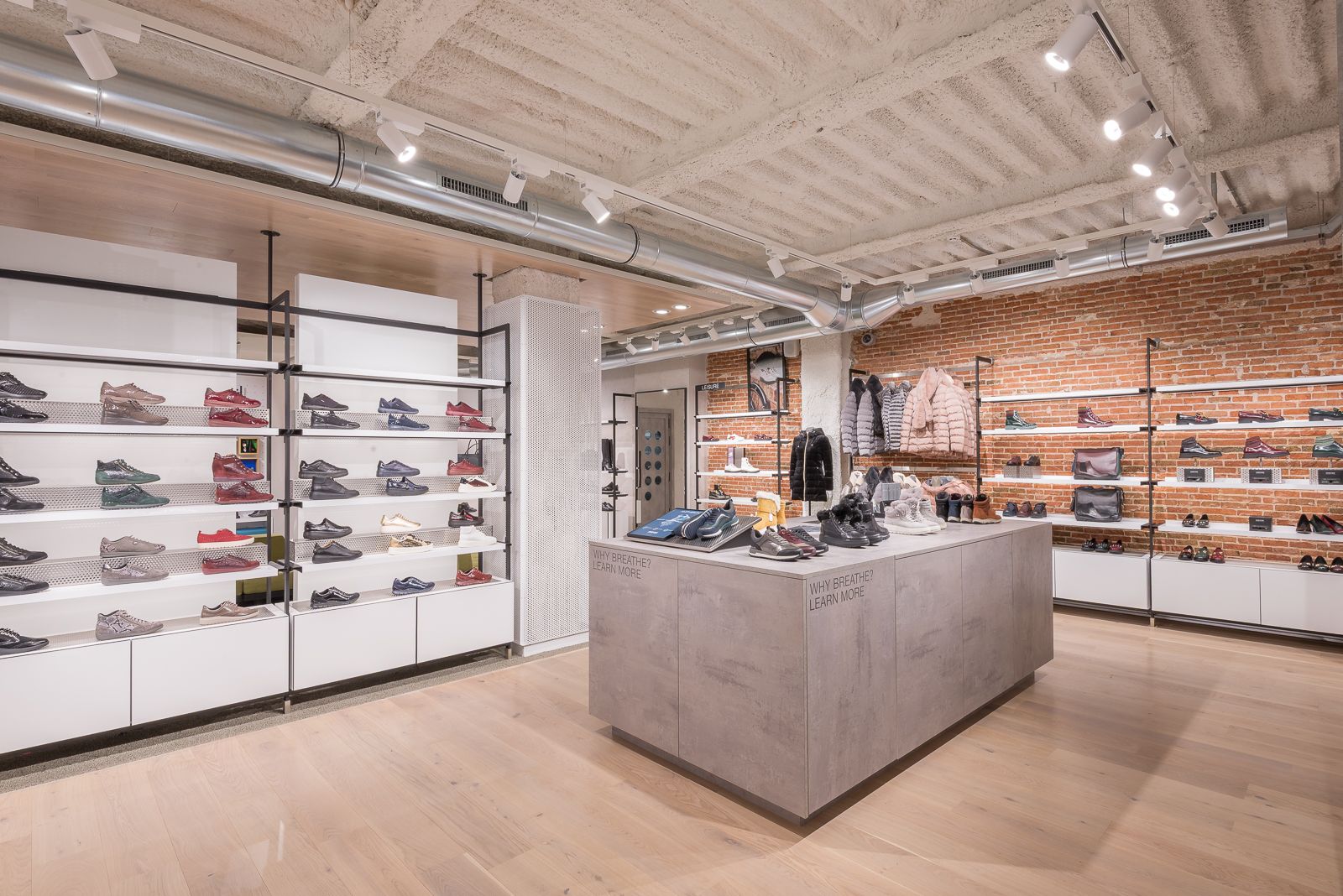 Interior de la tienda Geox, exhibiendo zapatillas coloridas en estantes y varias chaquetas de invierno en percheros contra una pared de ladrillo expuesto, bajo iluminación moderna y conductos expuestos.
