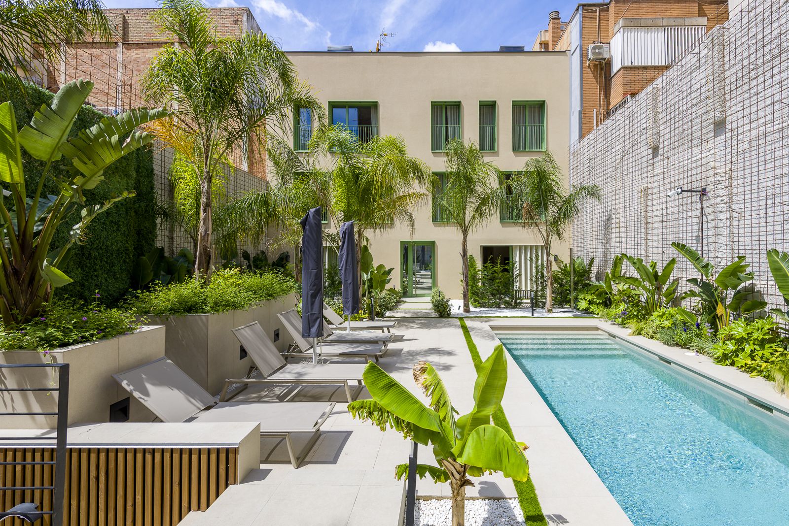 Bright outdoor courtyard featuring a clear blue lap pool, beige stone patio with sun loungers and umbrellas, and lush tropical greenery including palm trees and banana plants, set against a modern three-story building exterior with green accents.