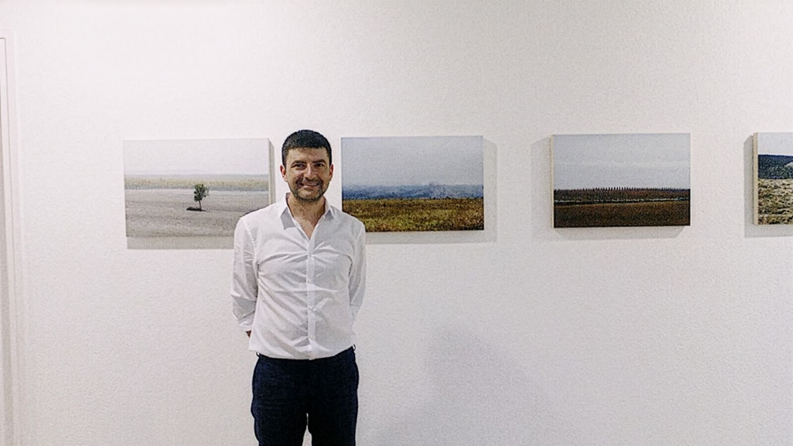 A man in a white shirt standing in front of landscape photographs on a gallery wall. The photographs depict various natural scenes, including fields and distant horizons.